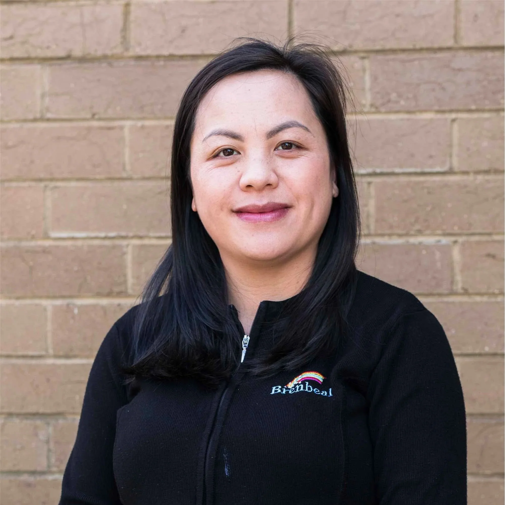 Close-up of a woman with black hair, wearing a black jacket with a rainbow logo and the word "Brinbeal" on it, standing in front of a brick wall.