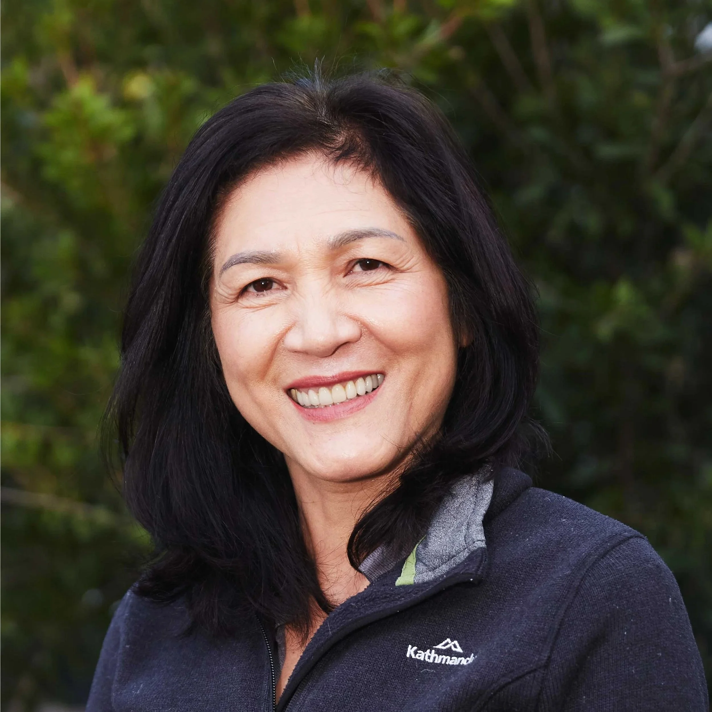 Close-up of an Asian woman with black hair and a broad smile, standing in front of a beige brick wall.