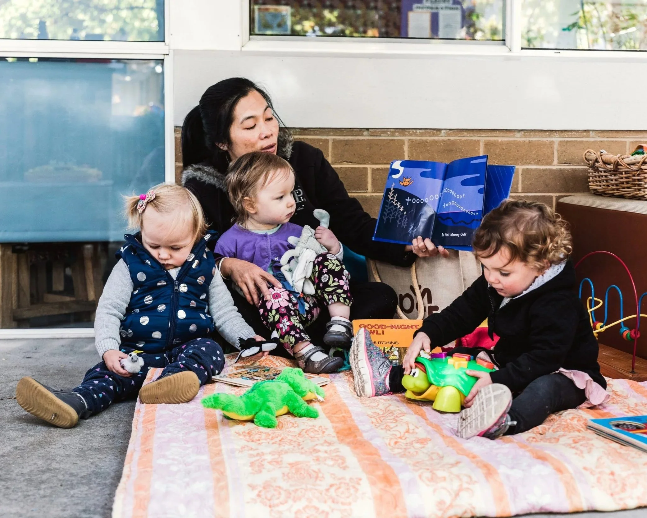 A woman with three young children playing indoors on a blanket with toys, reading a storybook, and holding a stuffed animal, against a background of a brick wall and large windows.