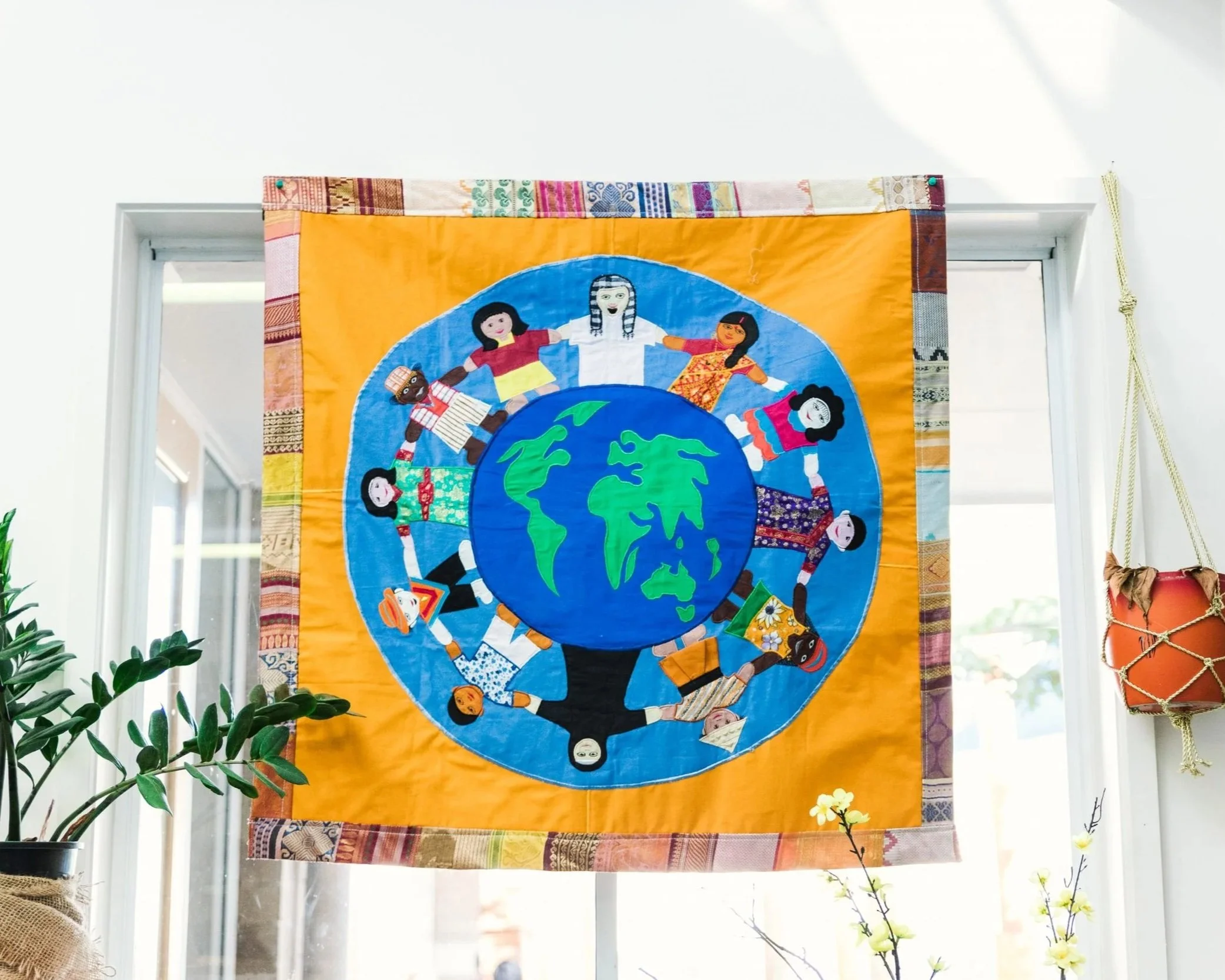 Colorful fabric art depicting diverse children holding hands around a globe on an orange background.