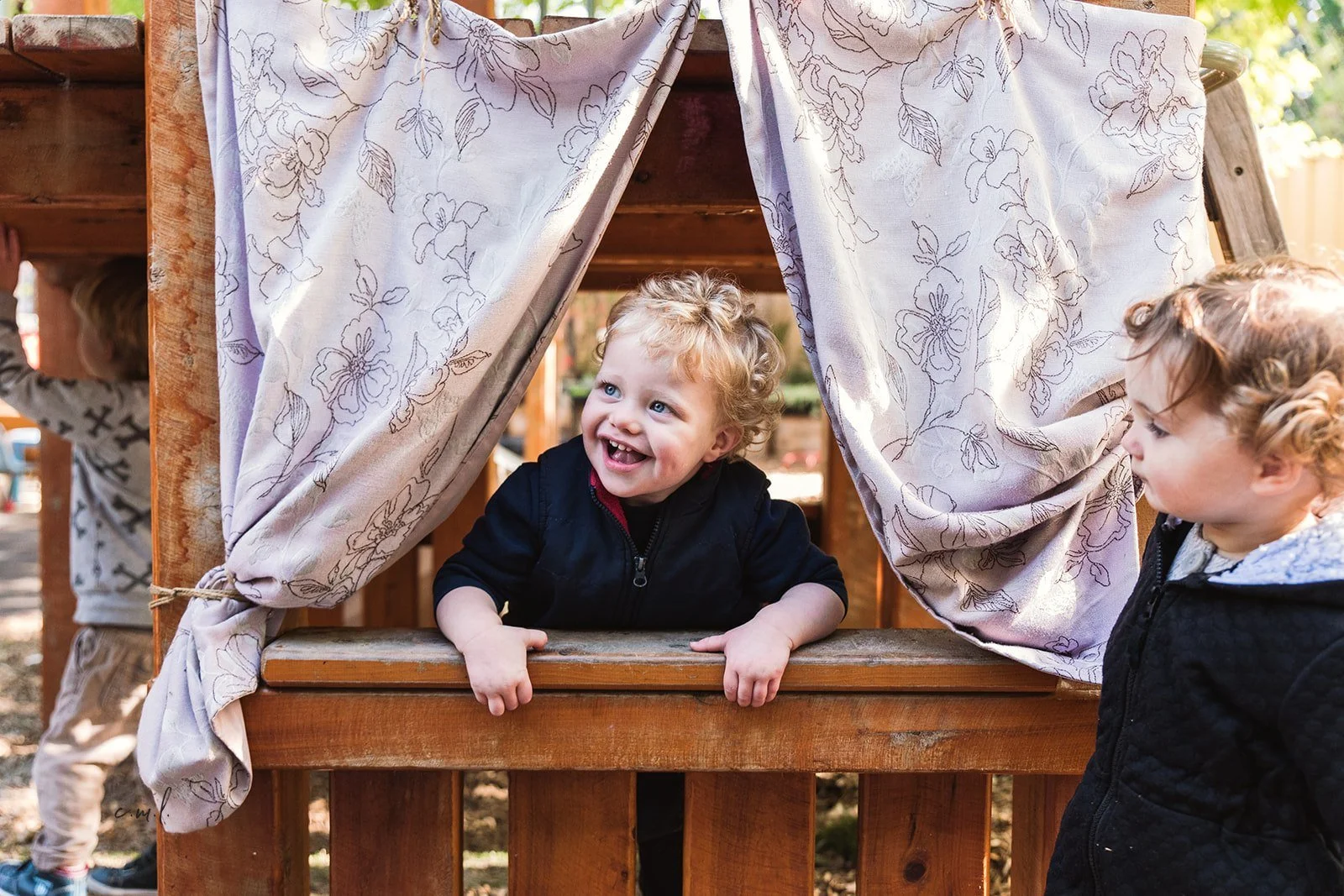 Two young children with curly hair interacting at a wooden playhouse. One child is inside the playhouse, leaning on the window ledge, smiling happily, while the other is standing outside, looking toward the child inside. The playhouse has light-colored curtains with floral patterns.