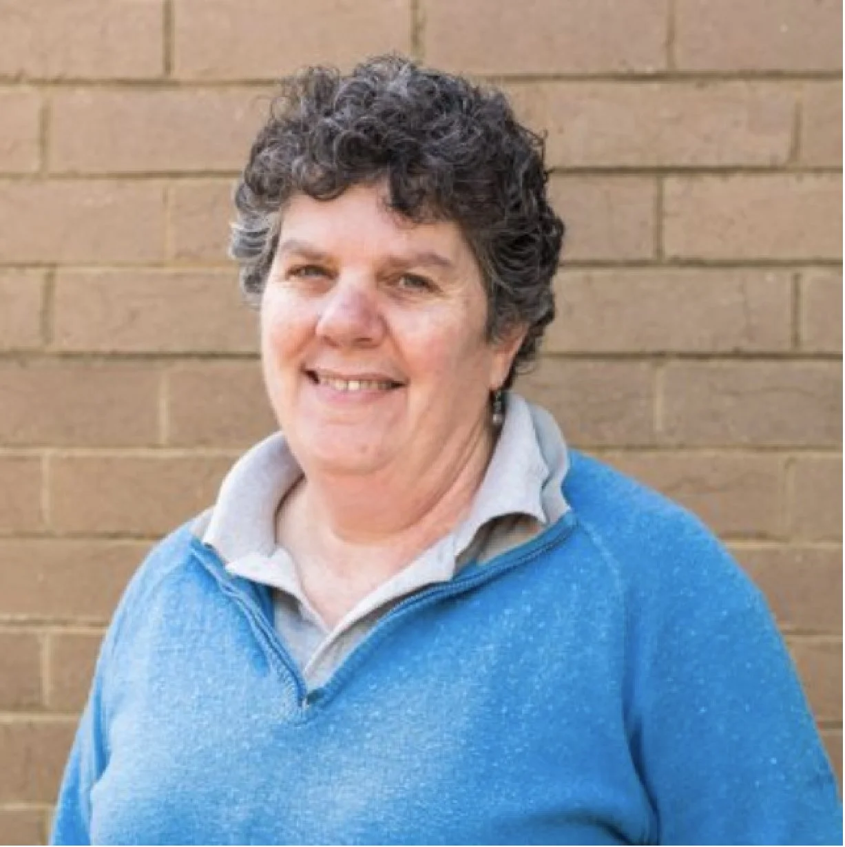 A smiling woman with short curly hair wearing a blue sweater and a gray collared shirt, standing in front of a brick wall.