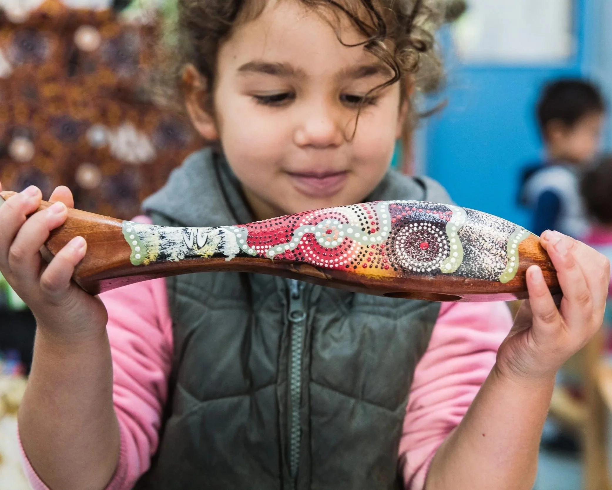 A young girl holding a wooden boomerang decorated with Aboriginal art patterns, looking at it curiously in an indoor setting.