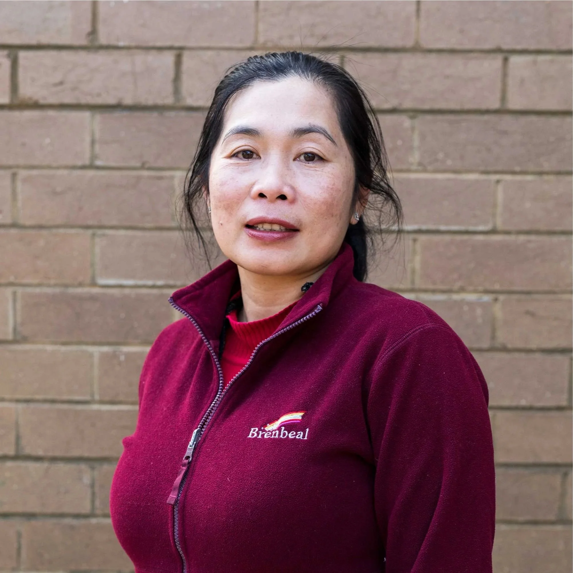 A woman with dark hair pulled back stands in front of a brick wall wearing a maroon fleece jacket with the word 'Brenbeal' embroidered on it.