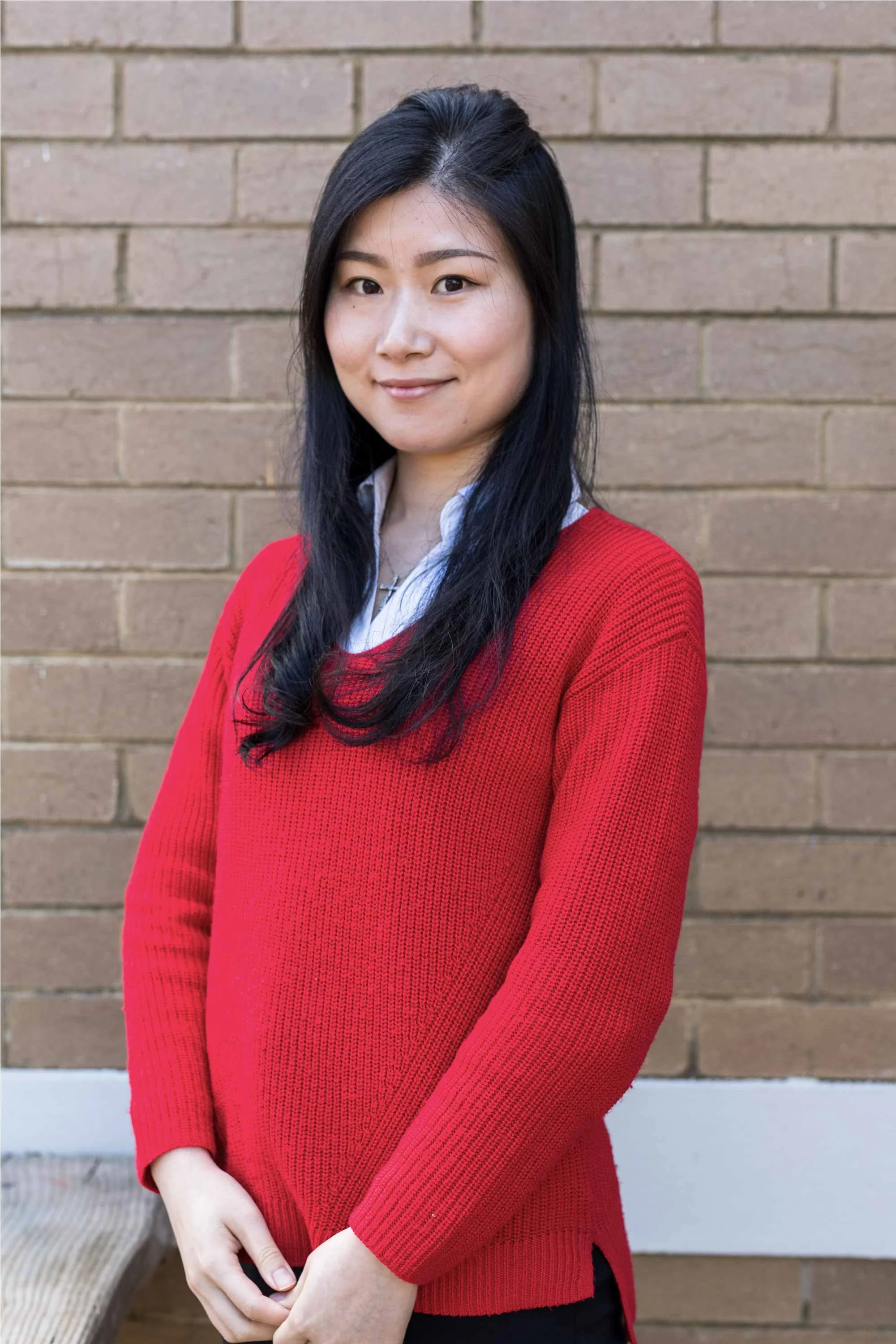 A woman with long black hair smiling at the camera, wearing a red sweater and a white shirt, standing in front of a brick wall.