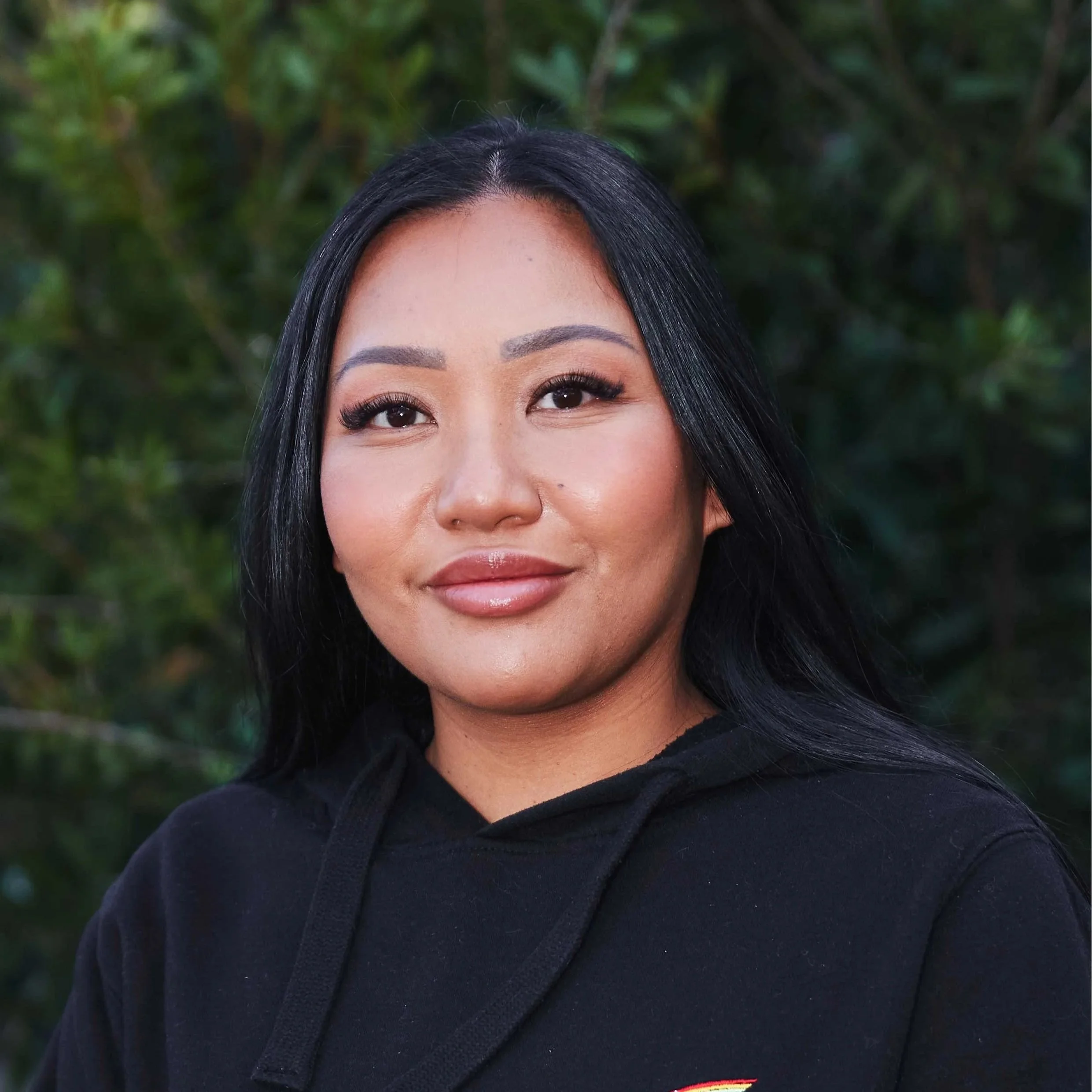 Close-up photo of a woman smiling with long black hair, wearing a black top, standing in front of a brick wall.