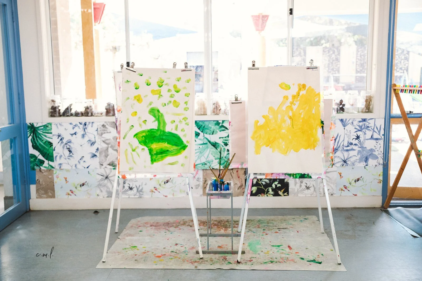 Two large abstract paintings on easels in a sunny room with windows, and a small table with painting supplies between them. The left painting has green shapes on a white background, and the right painting has yellow shapes on a light background.