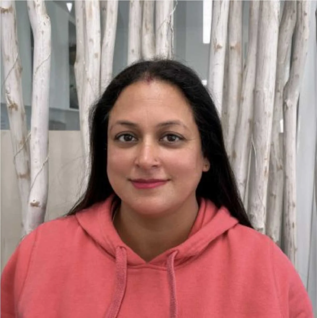 A woman with dark hair wearing a pink hoodie, smiling, standing in front of a background with white, textured tree branches.