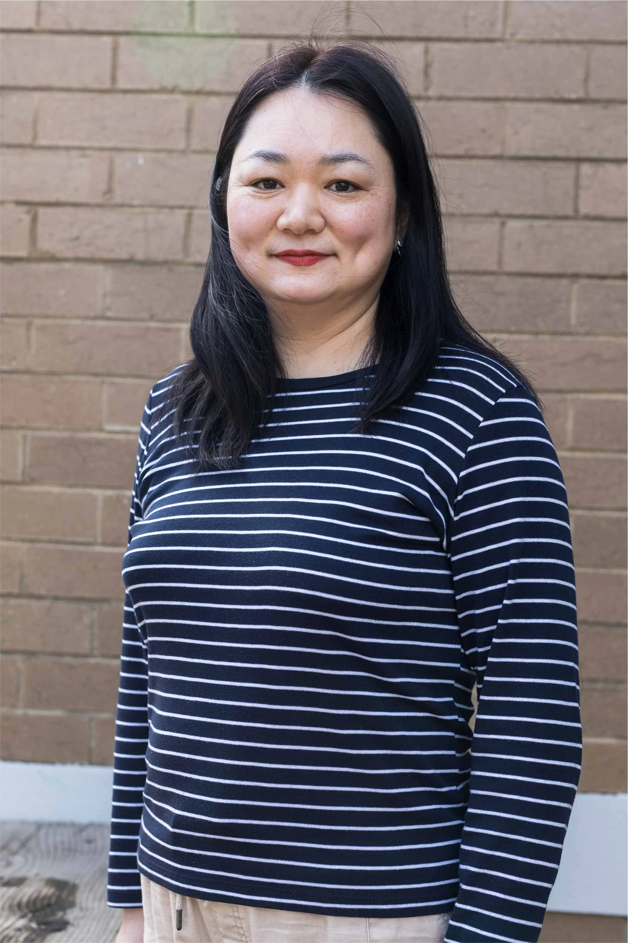 A woman with dark hair wearing a navy blue and white striped long-sleeve shirt standing against a brick wall.