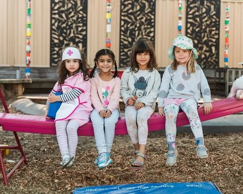 Four young girls sitting on a pink slide in a backyard playground, dressed in colorful clothing and hats, smiling at the camera.