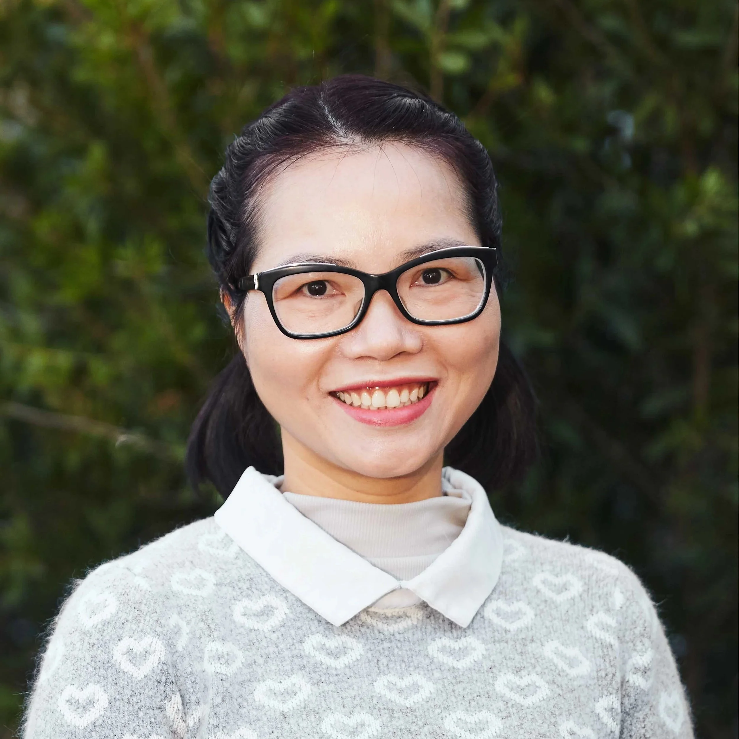 Close-up of a smiling woman with black hair, glasses, wearing a light pink sweater with lace details at the collar, standing in front of a brick wall.