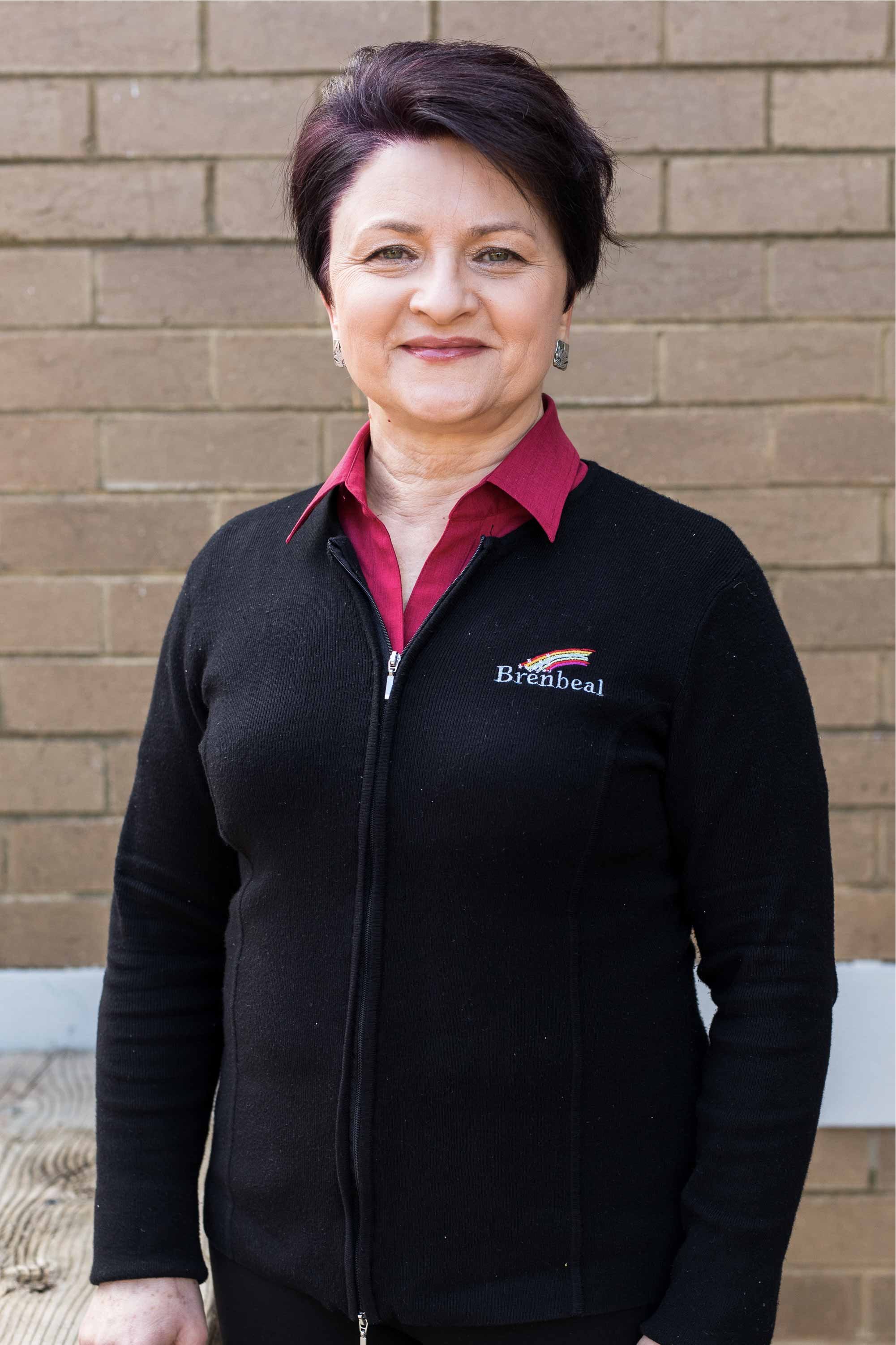 A middle-aged woman with short dark hair, wearing a black zip-up jacket with a logo and a dark red shirt underneath, standing in front of a brick wall.