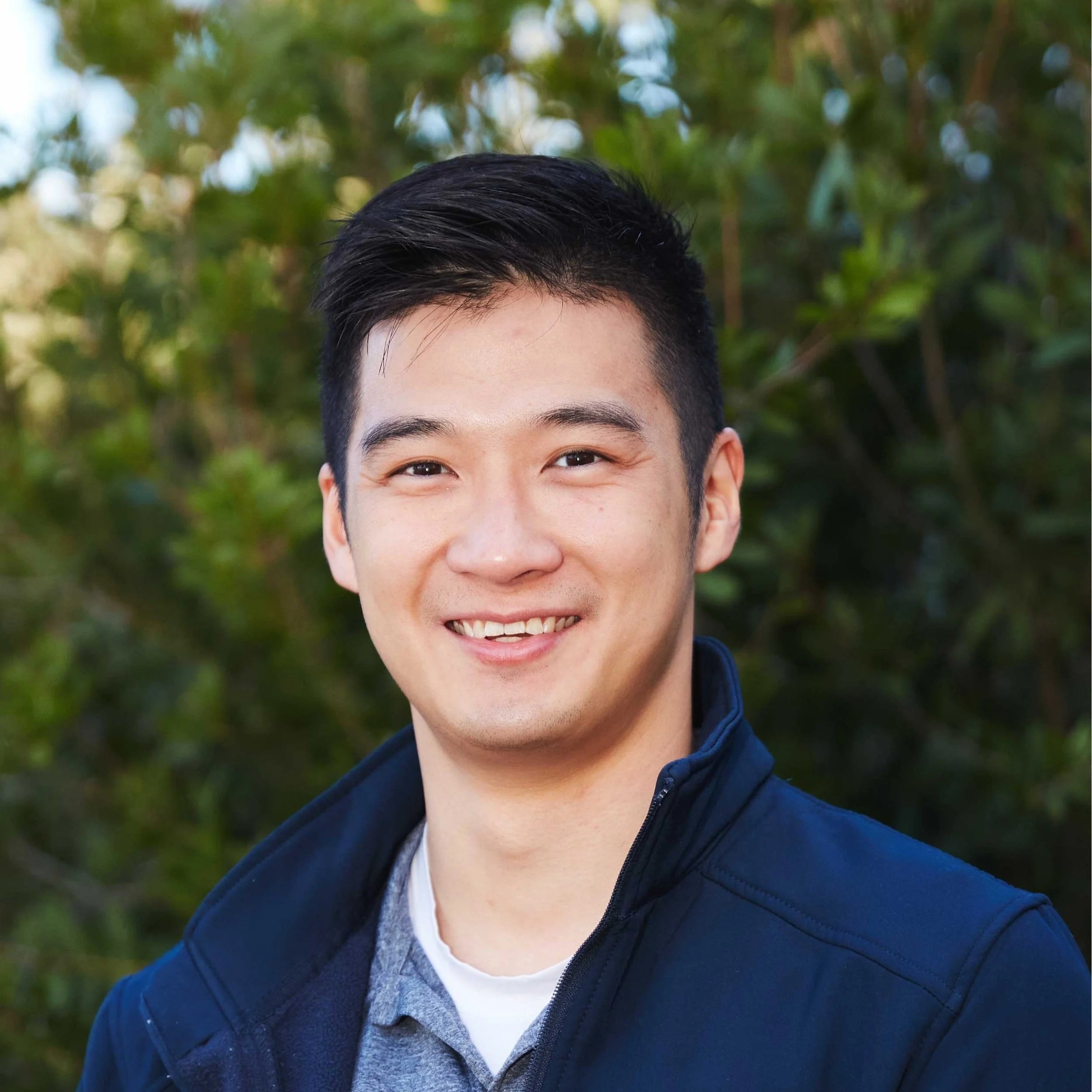 A young man smiling outdoors with a blurred green foliage background.