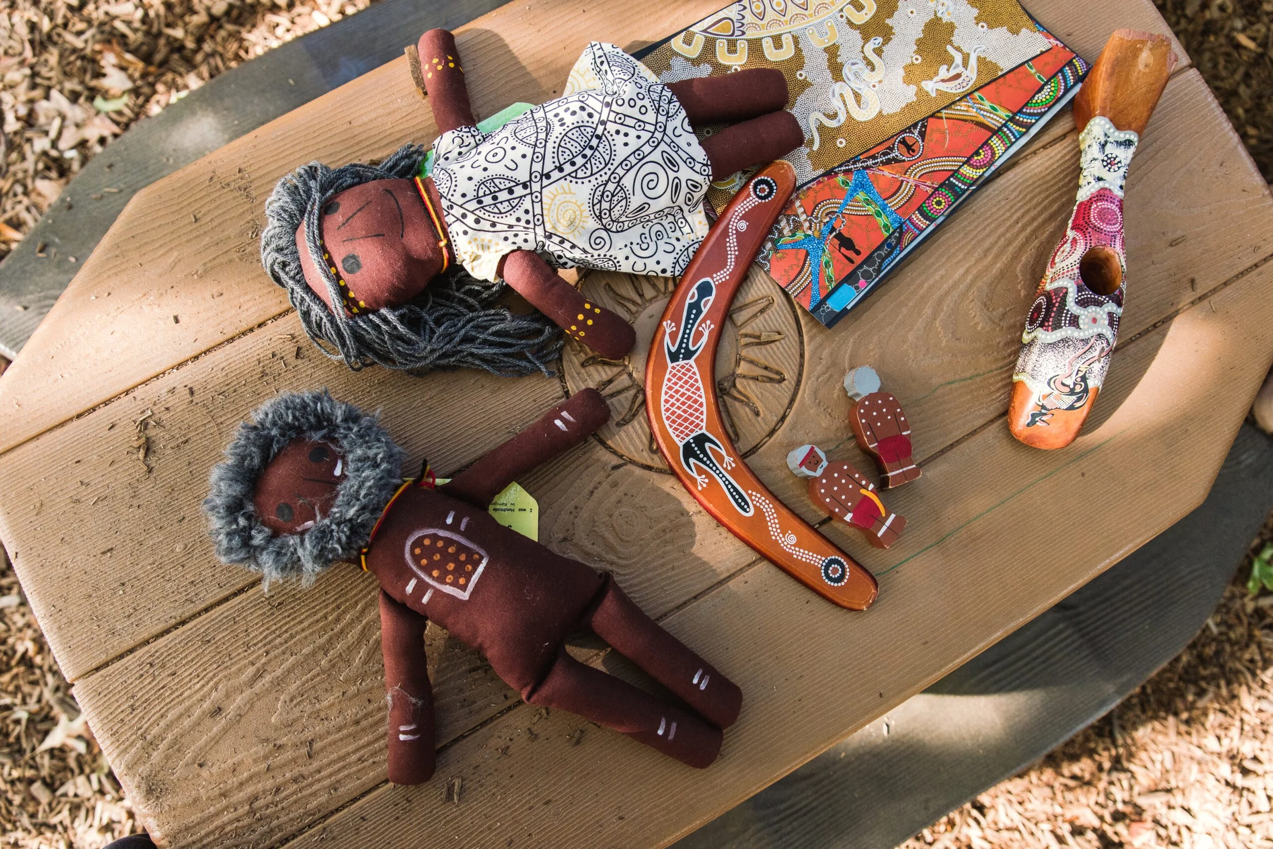 A wooden outdoor table displays various handcrafted Aboriginal art-inspired dolls and wooden animal figures, with a painted boomerang and a colorful book, surrounded by fallen leaves.