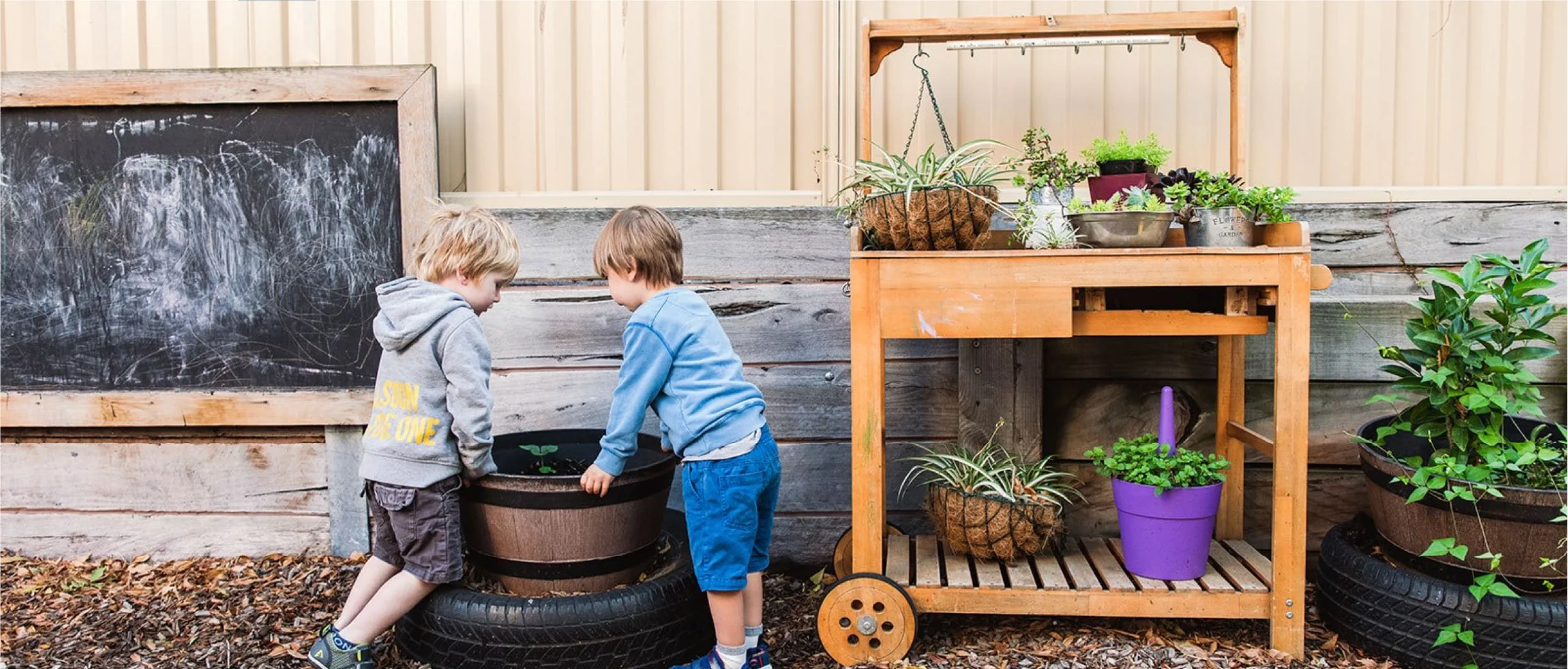 Two young boys with brown hair and casual clothing gardening with plants in pots outdoors, next to a chalkboard and a wooden gardening cart with potted plants.