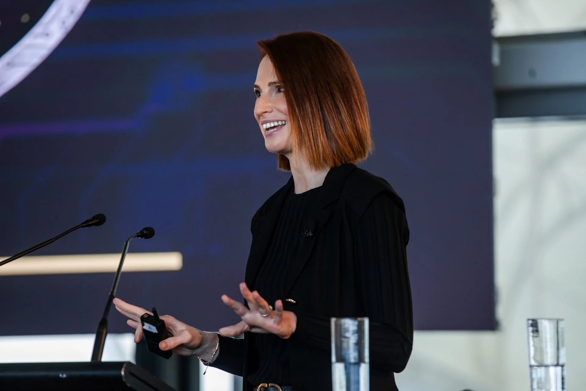 A woman with shoulder-length red hair is speaking at a podium with two microphones. She is wearing a black outfit and is smiling.