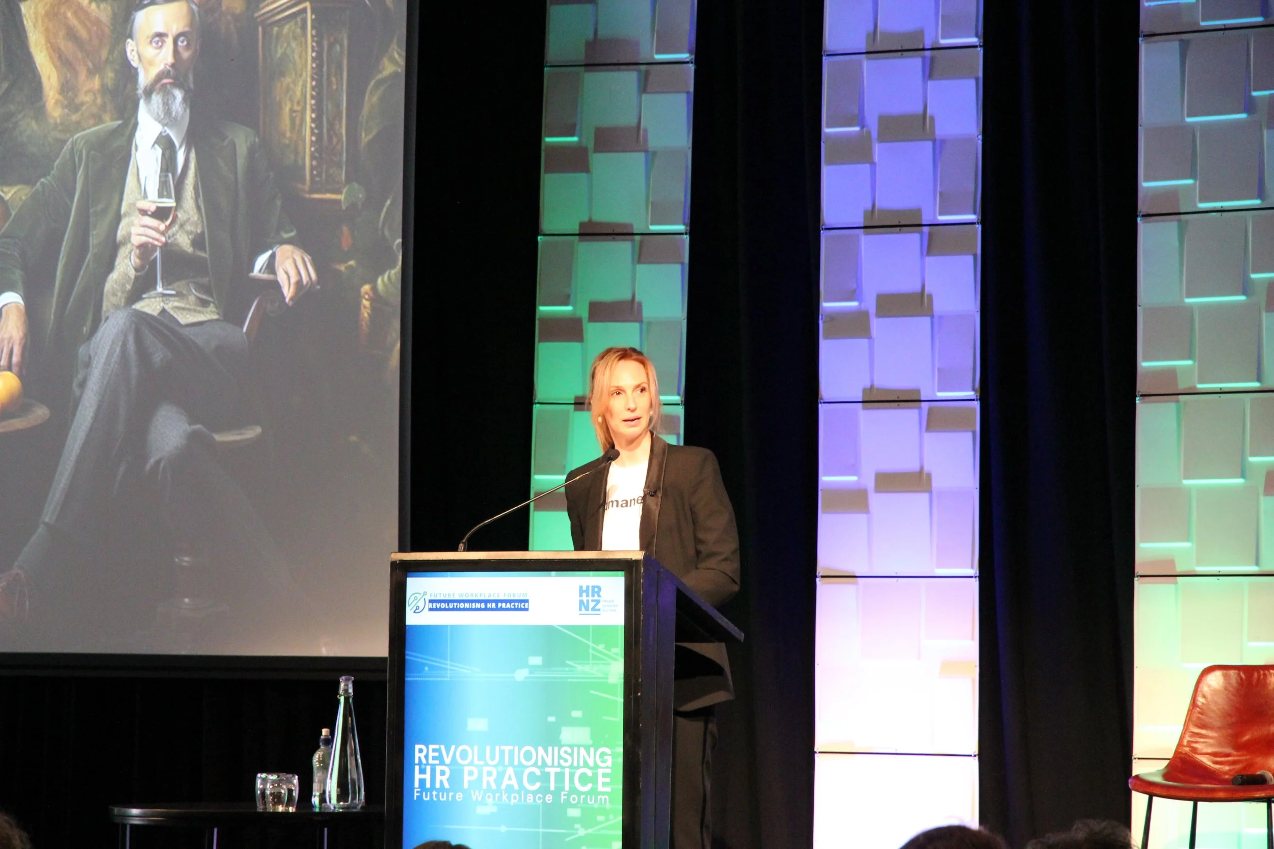 Annie Johnson speaking at a conference podium with a large screen behind her showing an artwork of a man holding a glass of wine. The setting is a professional event with colorful lighting on the backdrop.