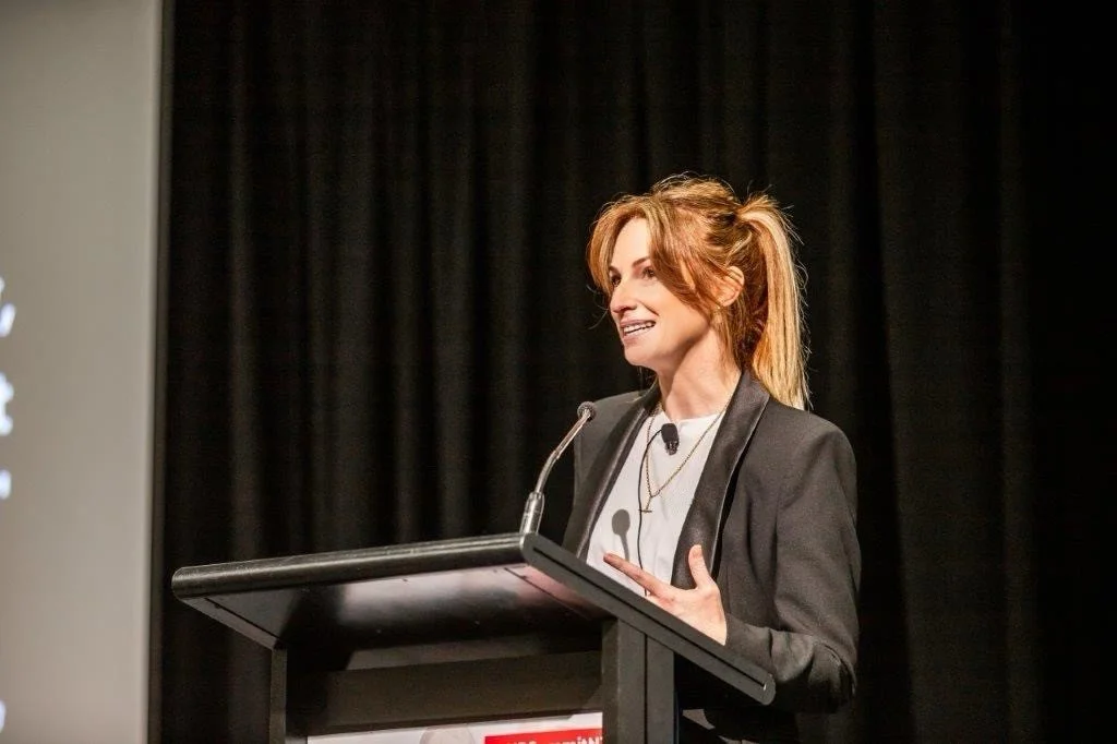 Annie Johnson standing at a podium, speaking into a microphone, with a dark curtain in the background.