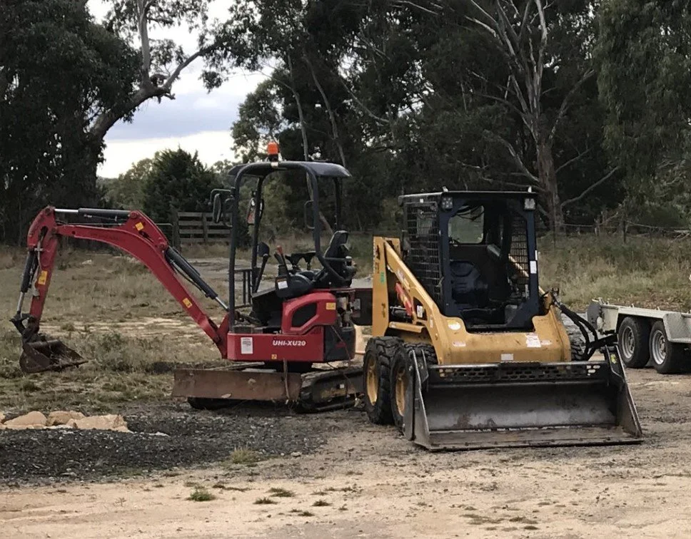 A small red excavator and a yellow skid steer loader are parked on a construction site with dirt, grass, and trees in the background.