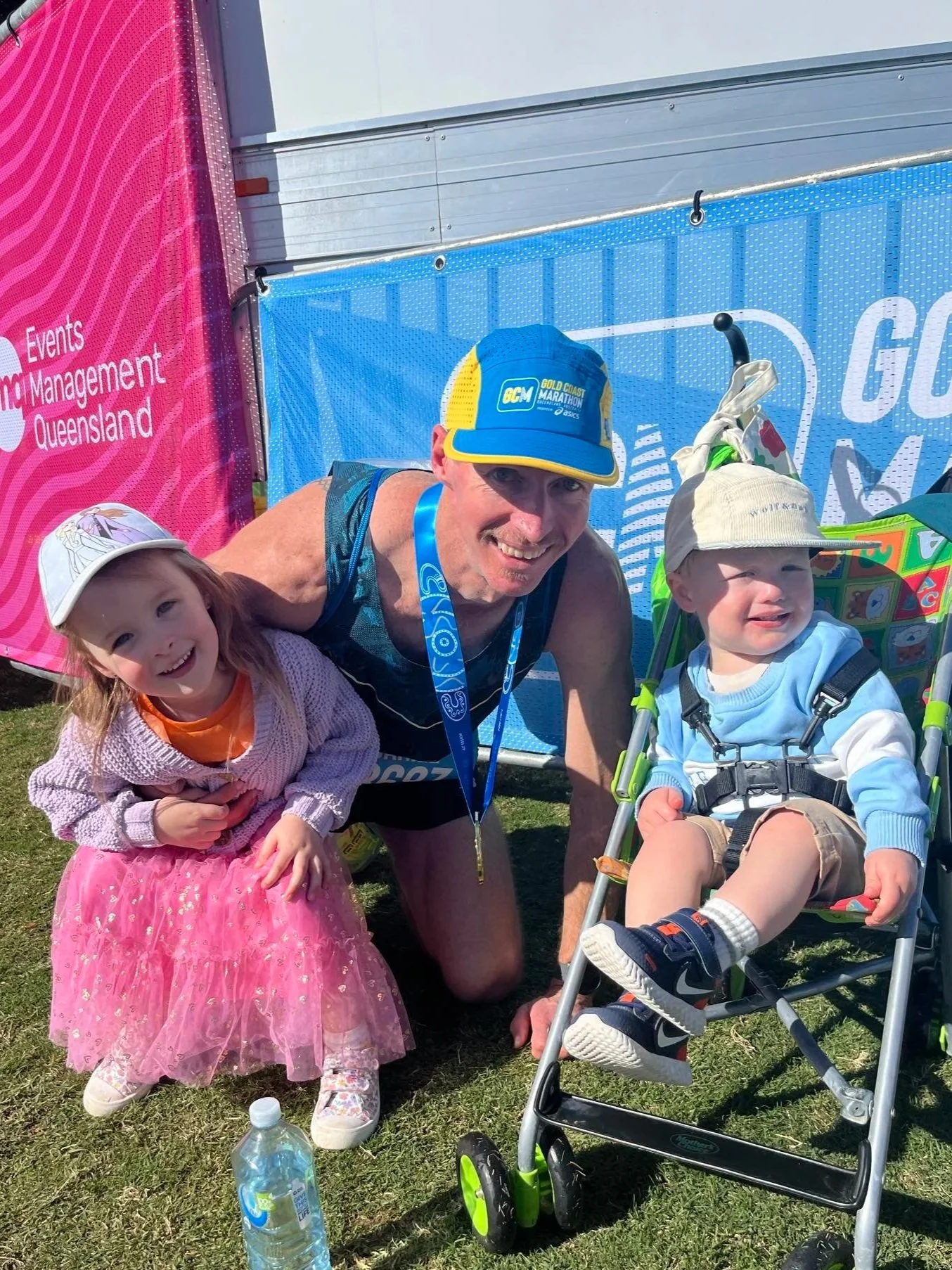 A man with two young children outdoors, smiling and posing for a photo after a marathon event. The girl wears a pink tutu and white hat, the boy is in a stroller with a beige hat, and there are event banners and a water bottle in the background.