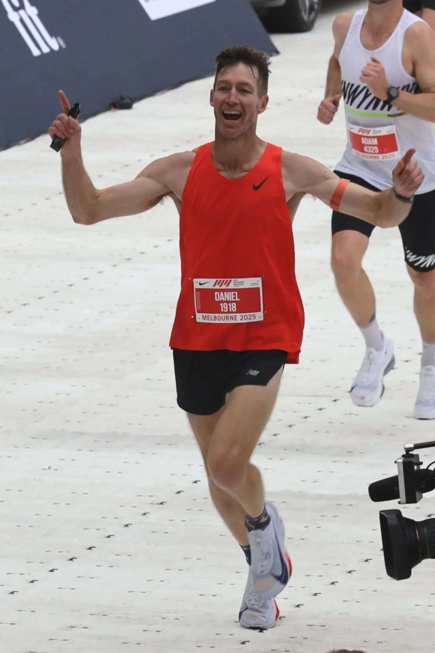 A male runner wearing a red Nike running singlet and black shorts, smiling and celebrating as he crosses the finish line in a race, with other runners nearby and a snowy ground.