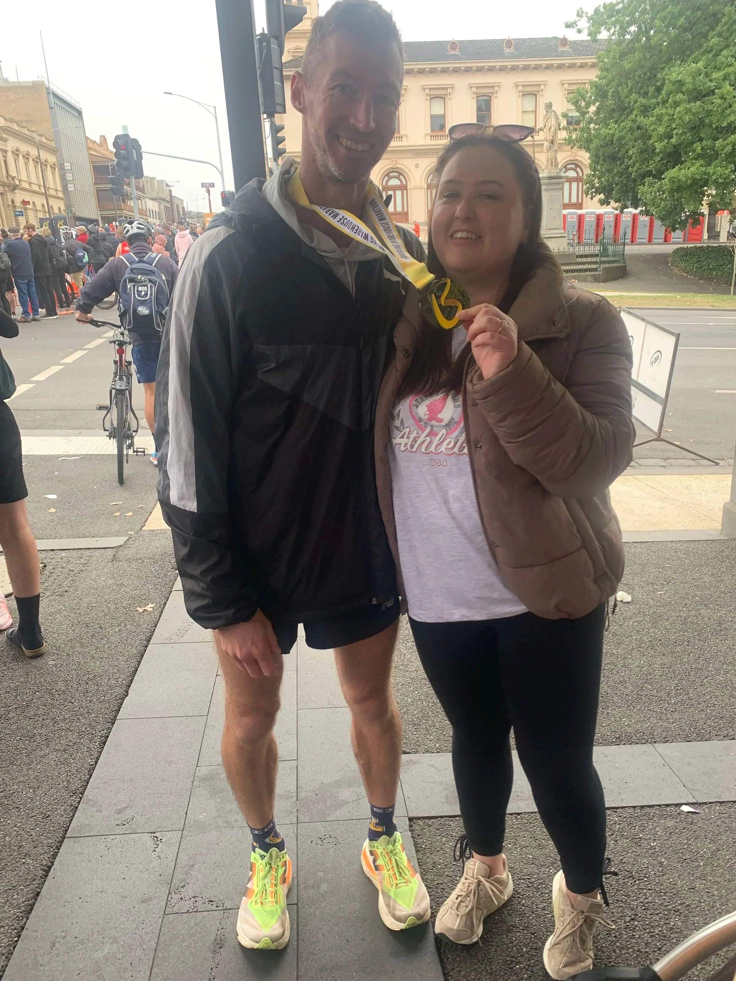 A man and woman standing together, smiling after a race, with medals around their necks, in an urban area with a crowd and historic buildings in the background.