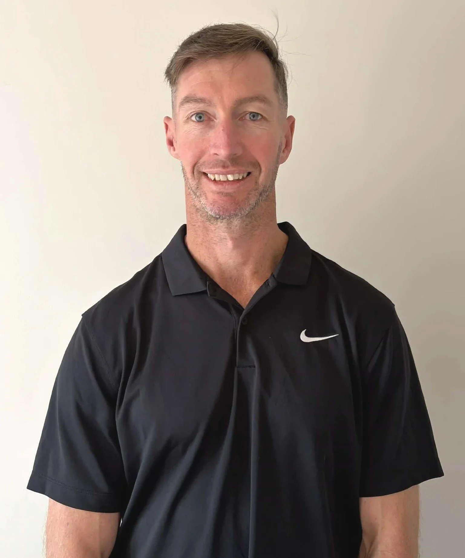 A smiling man with short brown hair and blue eyes, wearing a black Nike sports shirt, standing against a plain off-white wall.