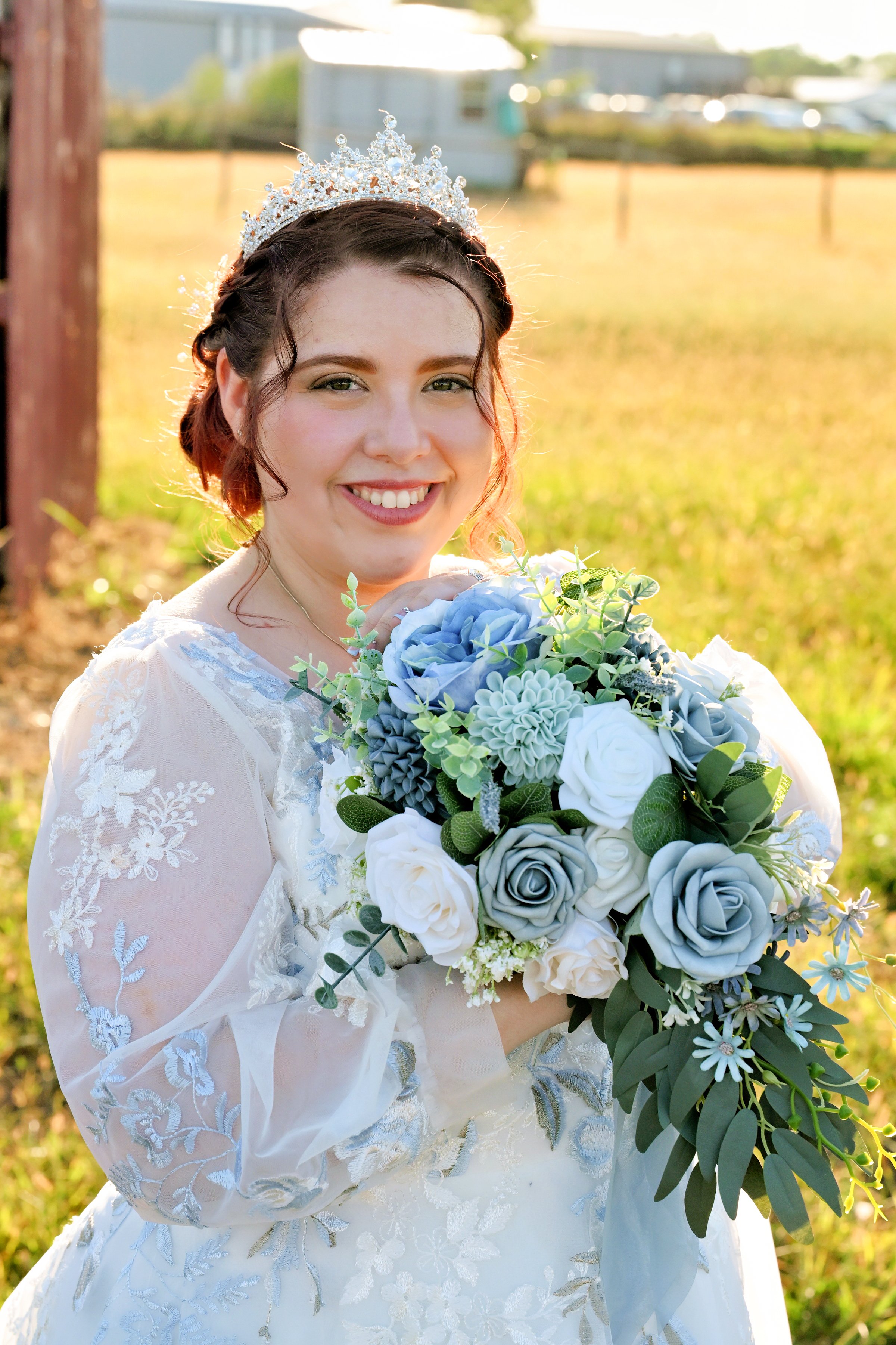 Bride in a white lace wedding gown with a tiara, holding a bouquet of white, gray, and blue flowers, smiling outdoors in a sunny field.