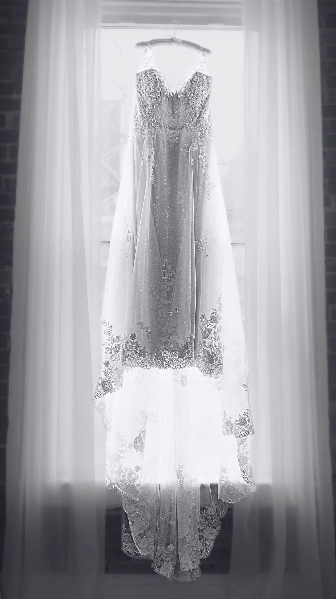 A wedding dress hanging on a hanger in front of a window with white curtains, backlit by natural light.