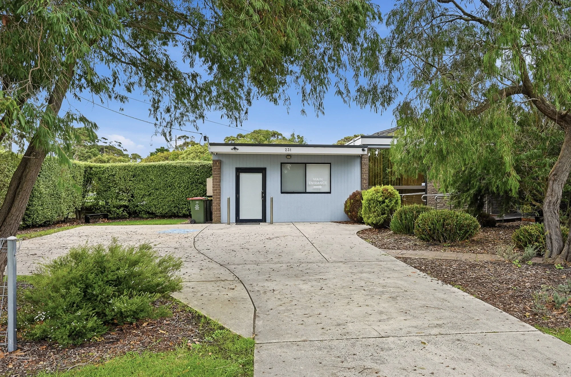 Front view of a small building with a sign that reads 'Main Entrance', surrounded by trees and shrubs, with a paved pathway leading to the door.