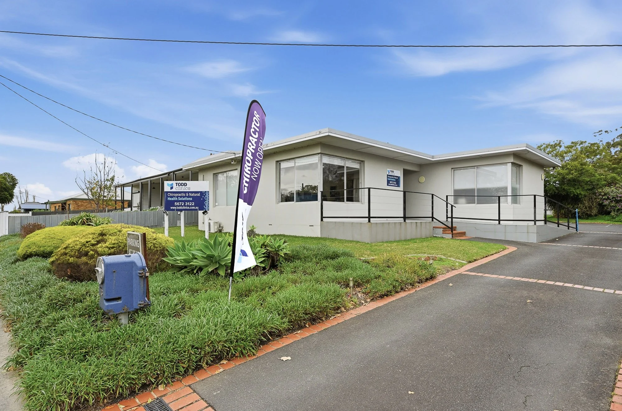 Outdoor view of a single-story chiropractic clinic building with large windows, a small ramp access, and a sign that reads "Chiropractic & Natural Health Solutions." In the foreground, a grassy area with plants and a mailbox. A vertical purple flag sign advertises chiropractic services. The sky is partly cloudy with power lines overhead.