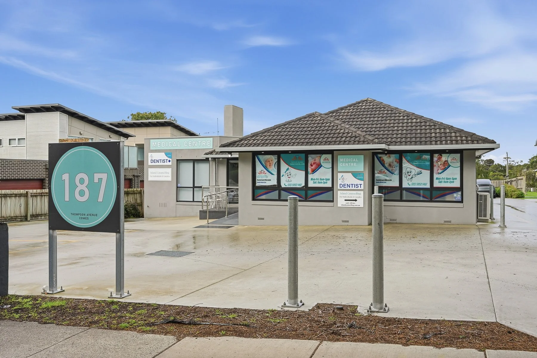 A small medical center building with glass windows and a roof with brown tiles. There are posters of children and dental symbols on the windows. In front, there is a sign with the number 187 in a teal circle, indicating an address or location, and the street name Thompson Avenue, Cowes. The parking area has poles and a small patch of dirt and grass along the sidewalk. The sky is clear with few clouds.