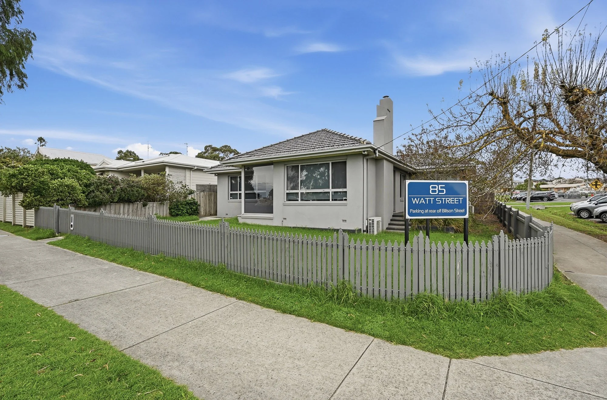 A suburban house with large front windows, a grey fence, a grassy yard, and a blue street sign indicating Watt Street. The street sign provides parking instructions for Bialson Street. There are trees, a parking lot, and neighboring houses in the background under a partly cloudy sky.