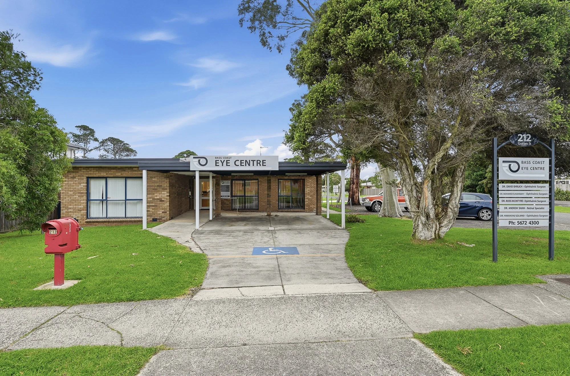 A small brick building with a sign reading 'Bass Coast Eye Centre' and an entrance, surrounded by trees and a grassy area. There is a red mailbox, a wheelchair accessible parking space, a sign listing ophthalmic surgeons, and cars parked in the background under a partly cloudy sky.