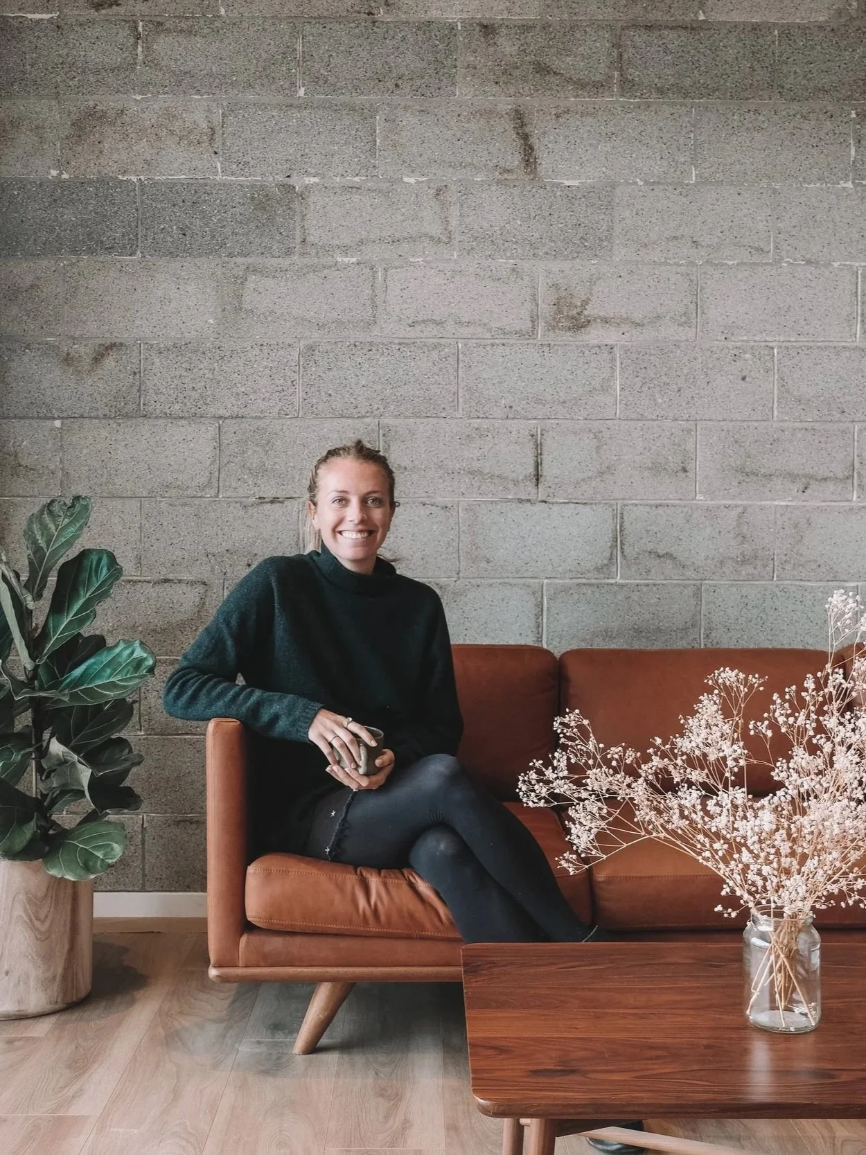 A woman sitting on a brown leather couch with a brick wall background, holding a coffee mug, with a large potted plant on her left and a glass vase with flowers on a wooden table in front of her.