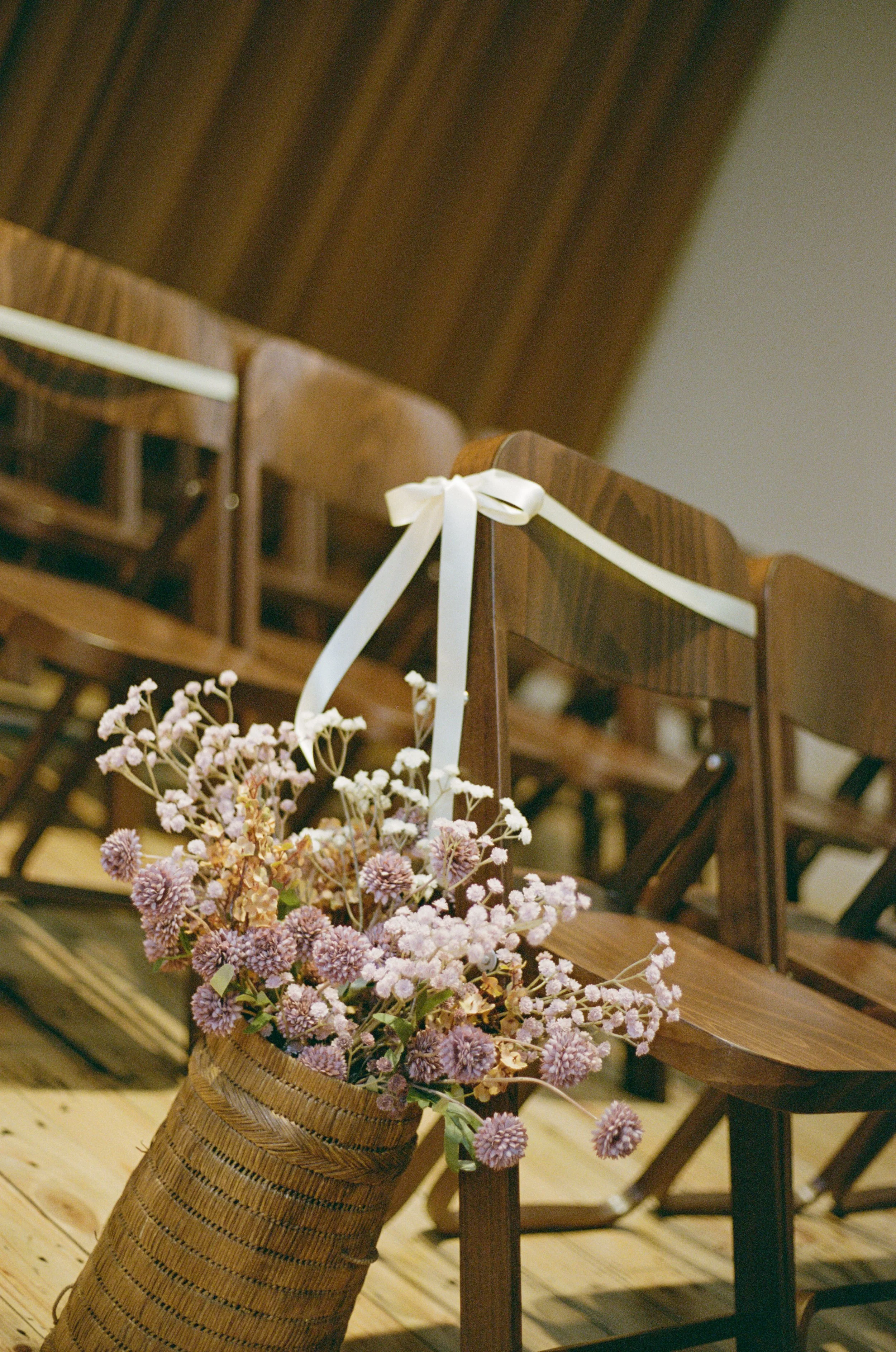 A bouquet of pink and purple flowers in a woven basket placed on a wooden floor, with wooden chairs in the background.