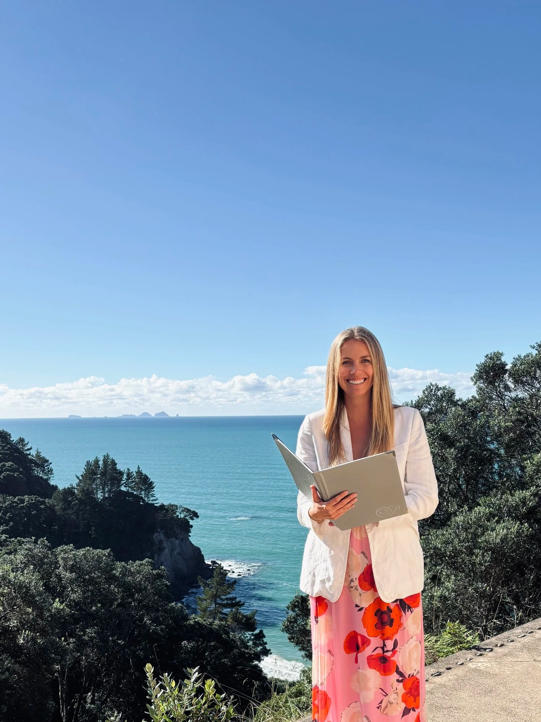 Young and modern wedding celebrant, based in Whangamata and serving venues in the Coromandel. Behind her is a scenic view of the ocean, distant islands, trees, and a clear blue sky.