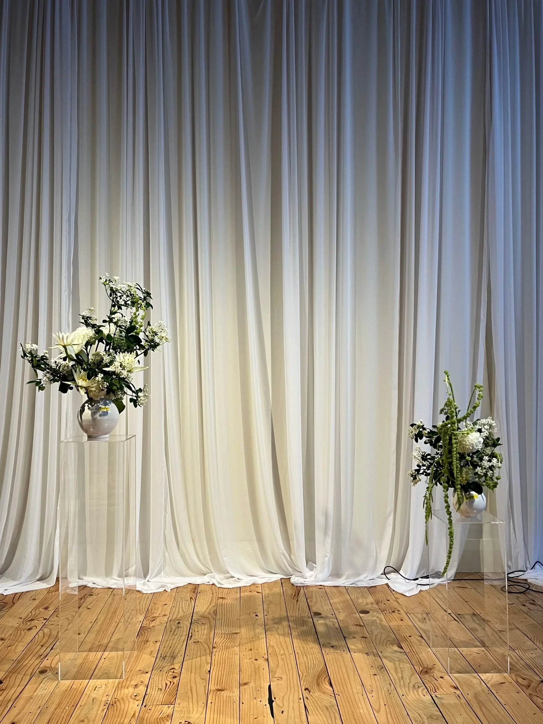 Two flower arrangements in white vases on clear stands in front of a white curtain backdrop with wooden floor.