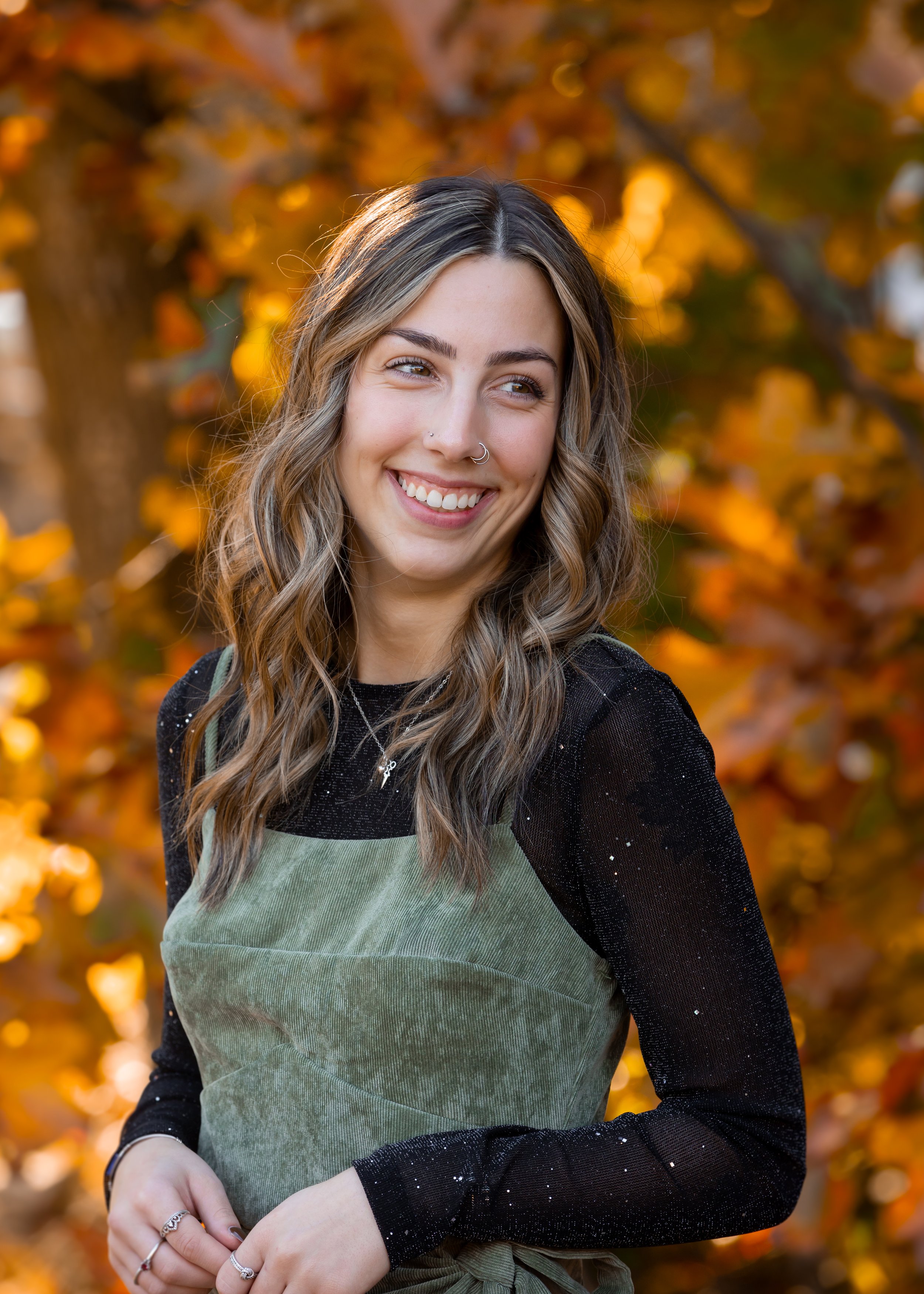 A woman with wavy brown hair and multiple nose and ear piercings smiling outdoors during autumn.