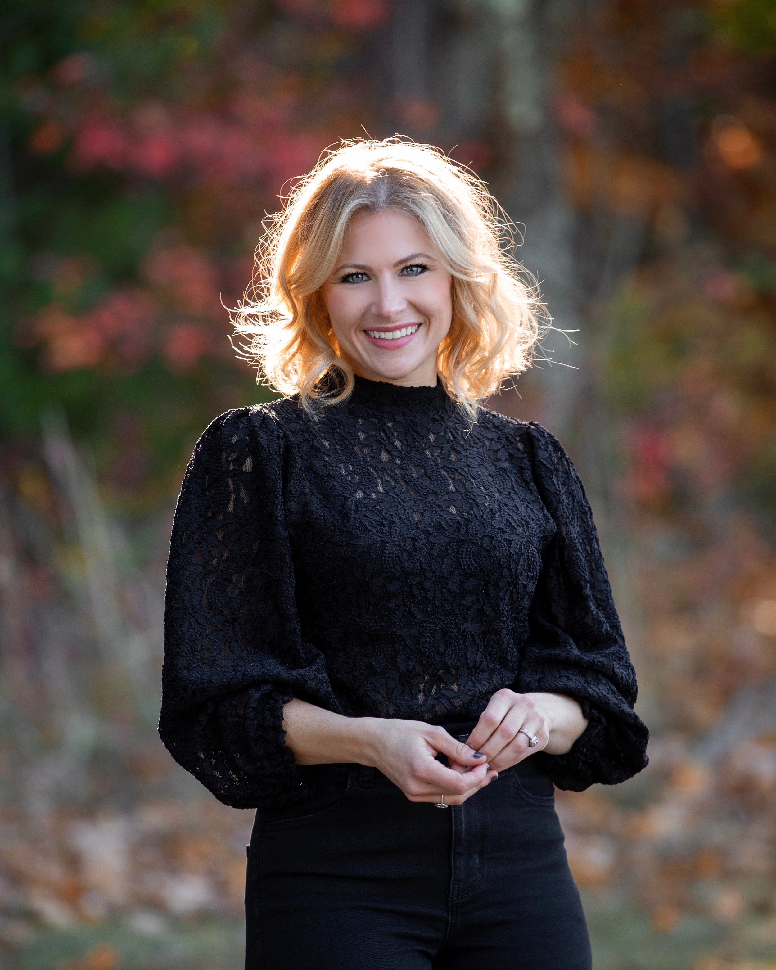 A woman with blonde hair, wearing a black lace top, smiling outdoors in front of blurred colorful autumn trees.