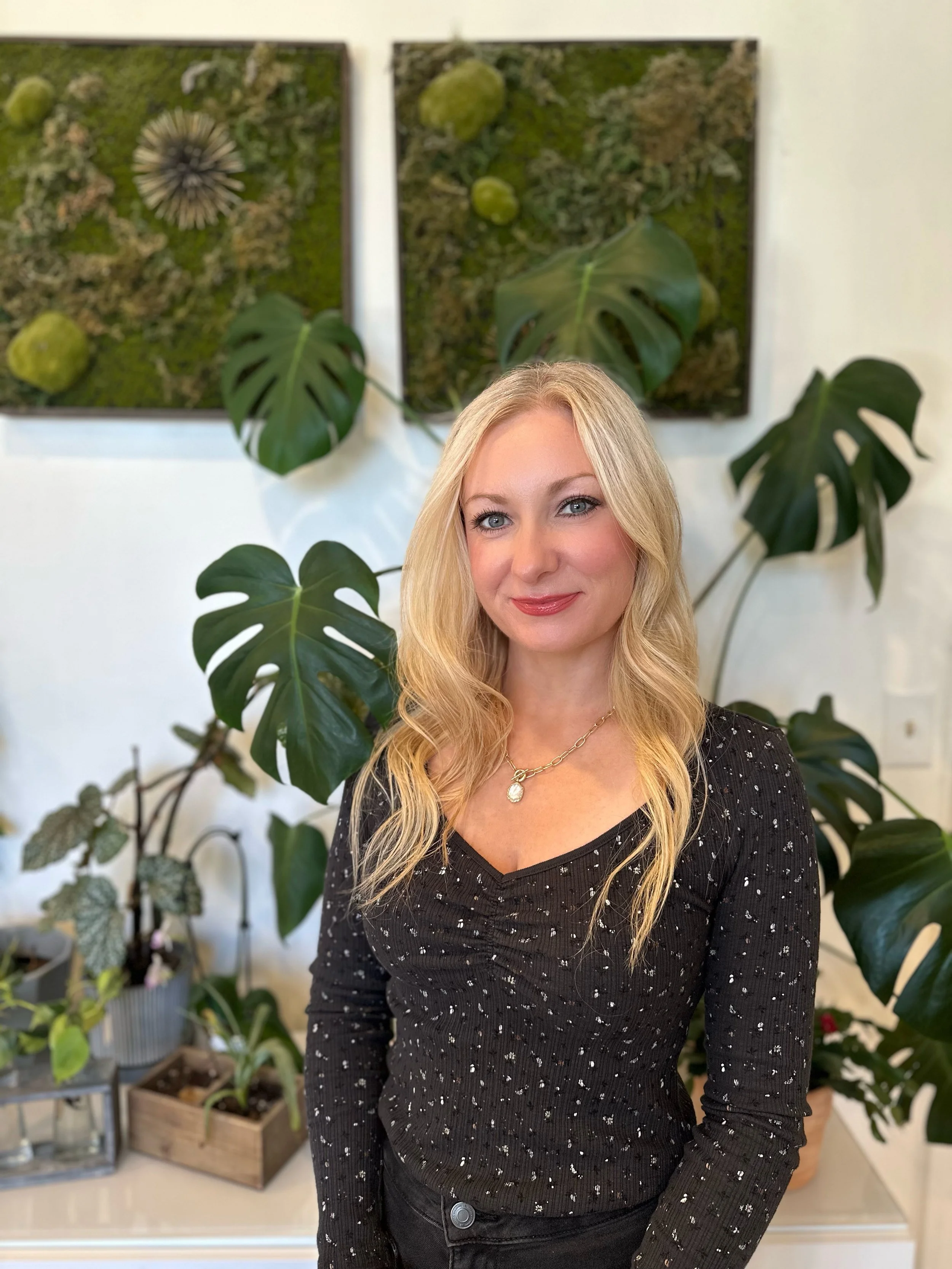 A woman with blonde hair and blue eyes, wearing a black sweater with white specks, standing in front of green plants and framed moss art on the wall.