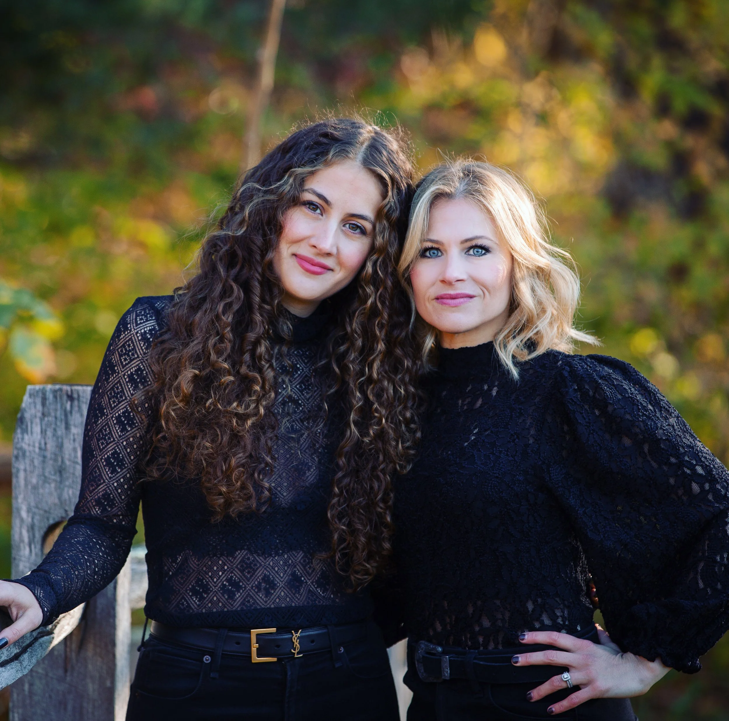 Close-up of two women standing outdoors in autumn, with blurred colorful fall foliage in the background. The woman on the left has curly brown hair and wears a black patterned top, while the woman on the right has wavy blonde hair and wears a dark blue textured sweater. Both women are smiling.