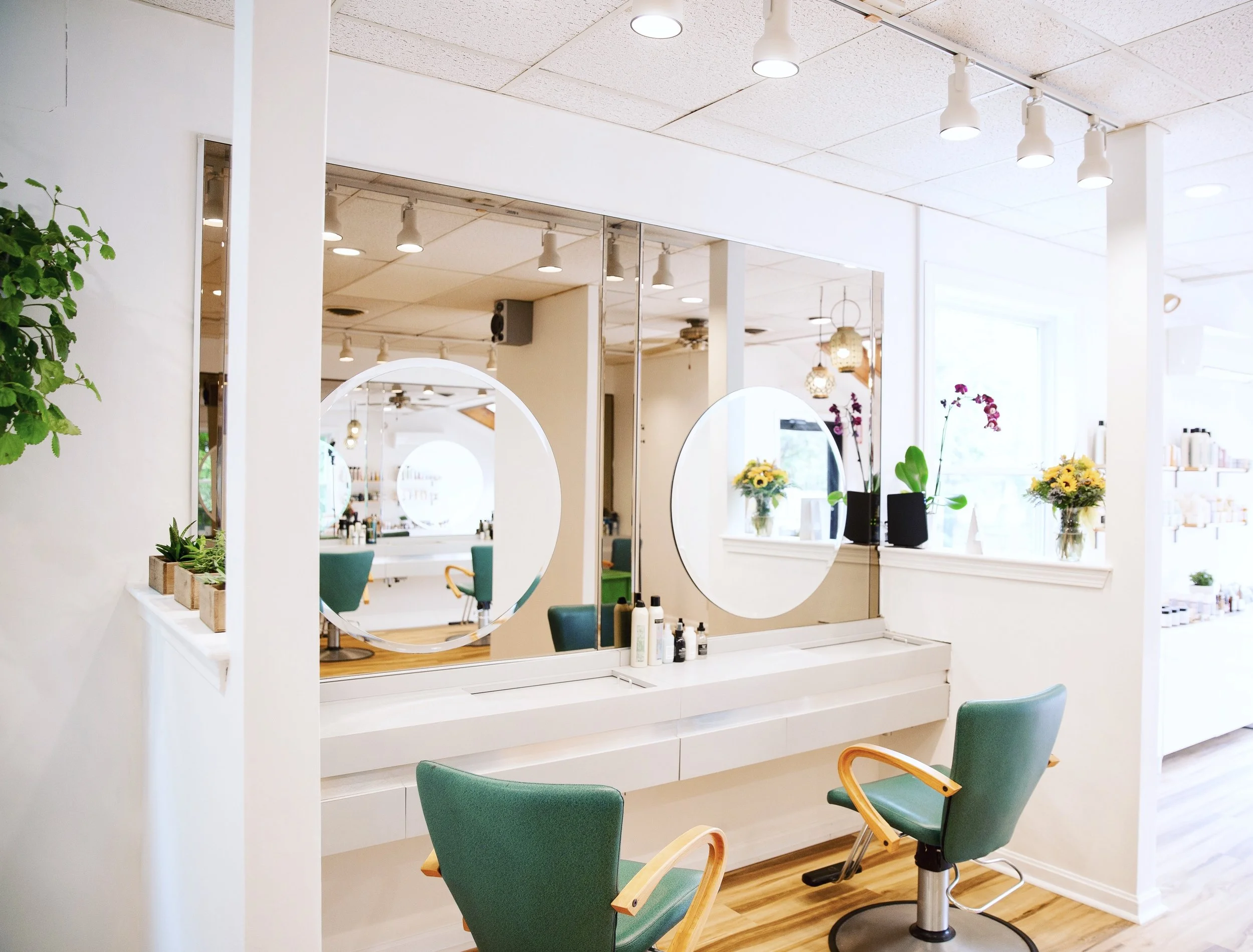 A modern hair salon interior with green chairs, large mirror, white walls, wooden flooring, and decorative plants and flowers, illuminated by ceiling lights.