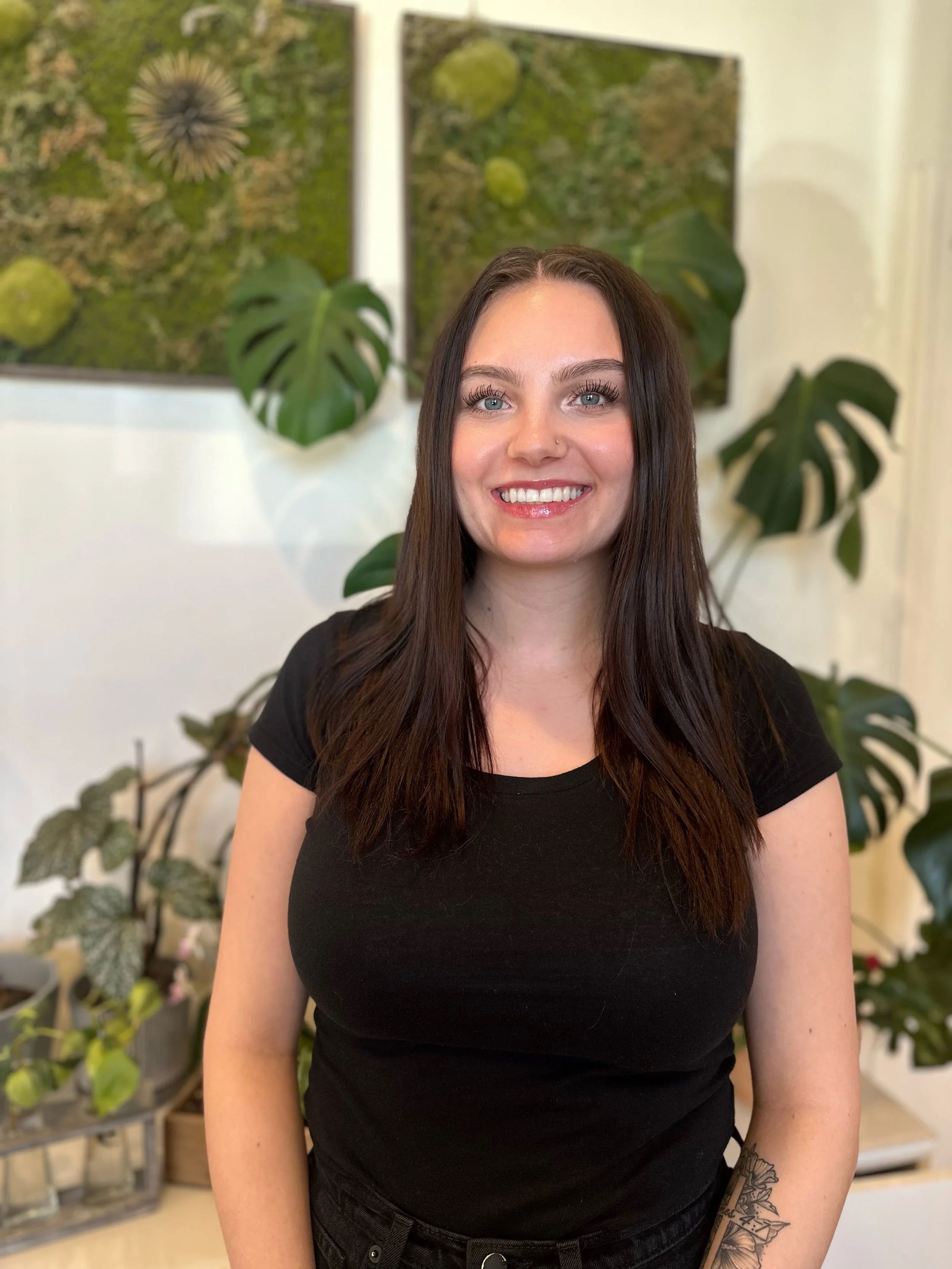 A woman with long dark hair smiling, wearing a black shirt, standing in front of a leafy green background with plants and a textured wall.