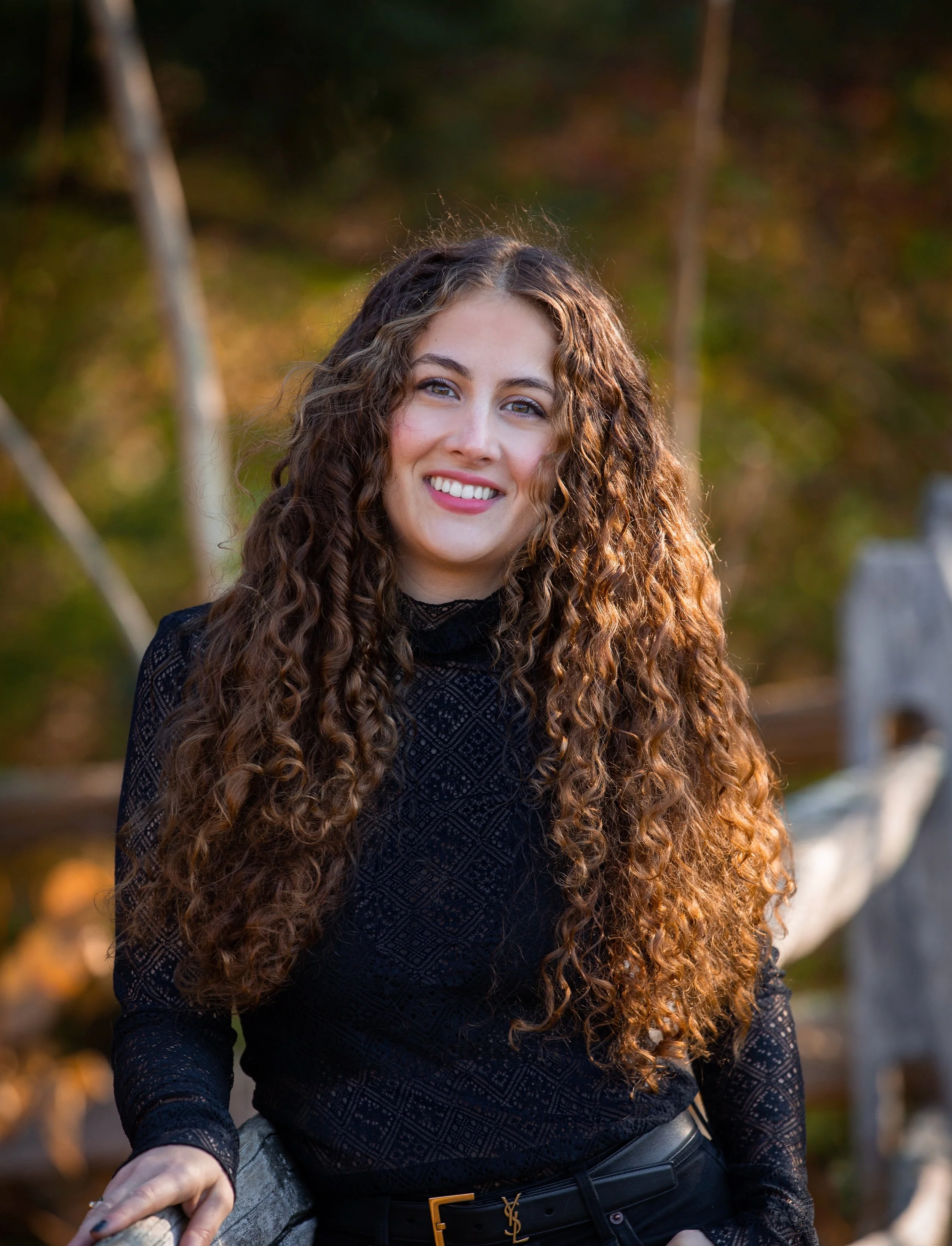 A woman with long curly hair, wearing a black lace top and a black belt with a gold YSL logo, stands outdoors with a smile on her face.