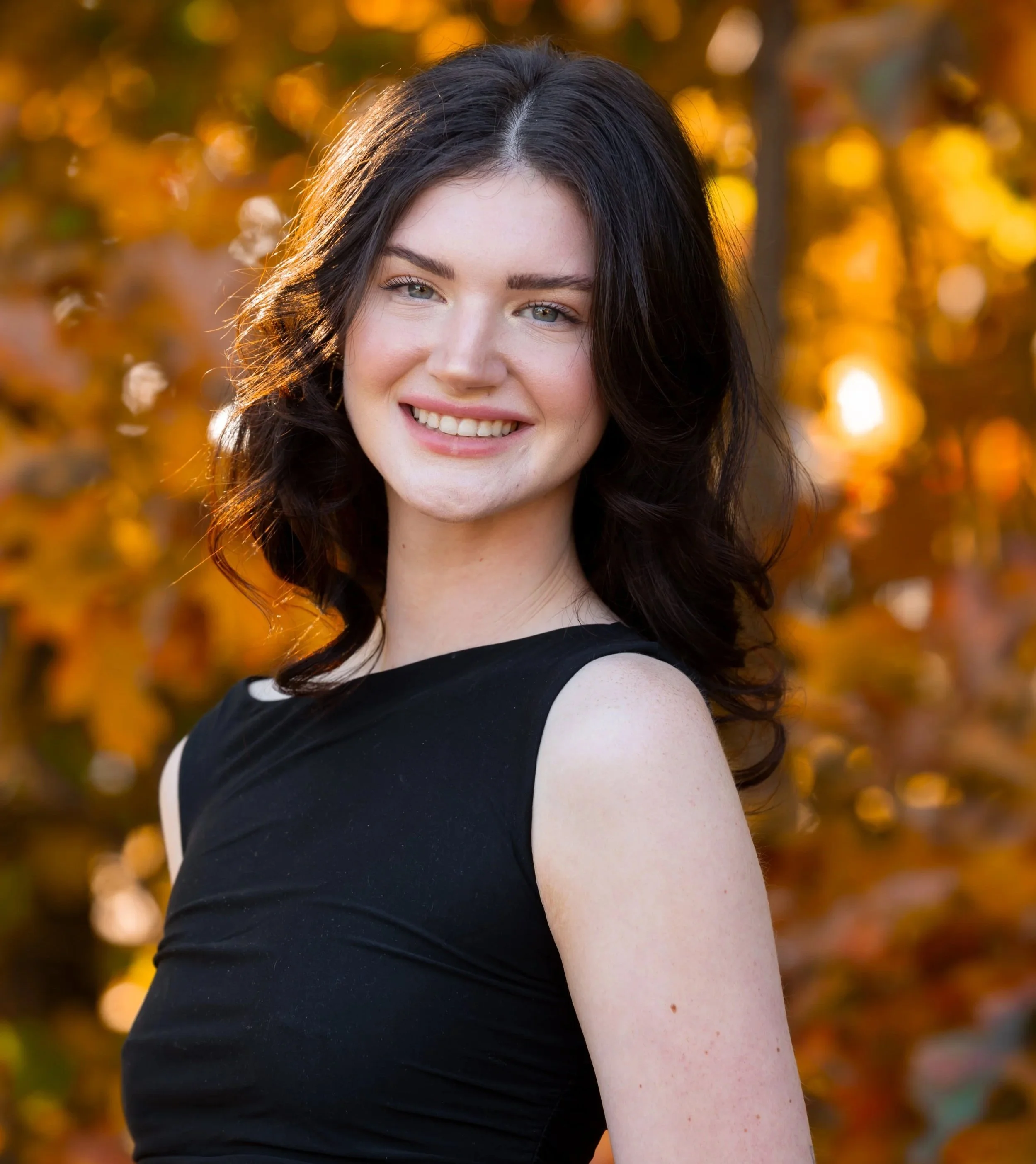 A smiling young woman with blue eyes and dark brown hair, standing outdoors in front of a background of blurred orange and yellow autumn leaves.