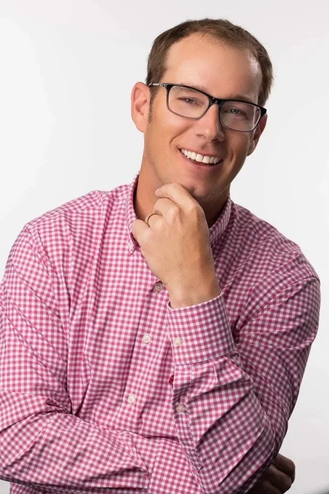 Young man with glasses wearing a pink checkered shirt, smiling, touching his chin with his hand.
