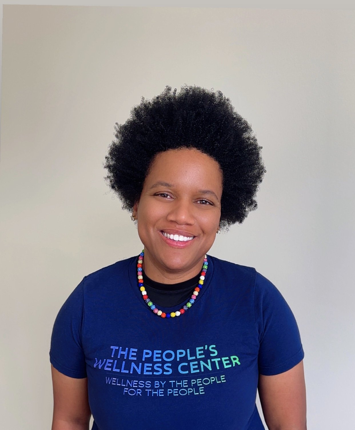 Smiling woman with natural afro hairstyle wearing a colorful beaded necklace and a navy blue T-shirt that reads "The People's Wellness Center" against a plain light-colored background.