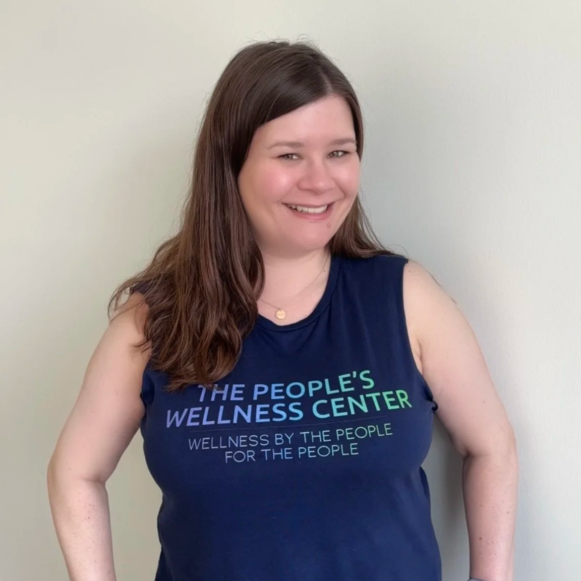 Smiling woman with brown hair wearing a navy blue sleeveless shirt that reads "The People's Wellness Center" and "Wellness by the people for the people" against a plain light-colored wall.