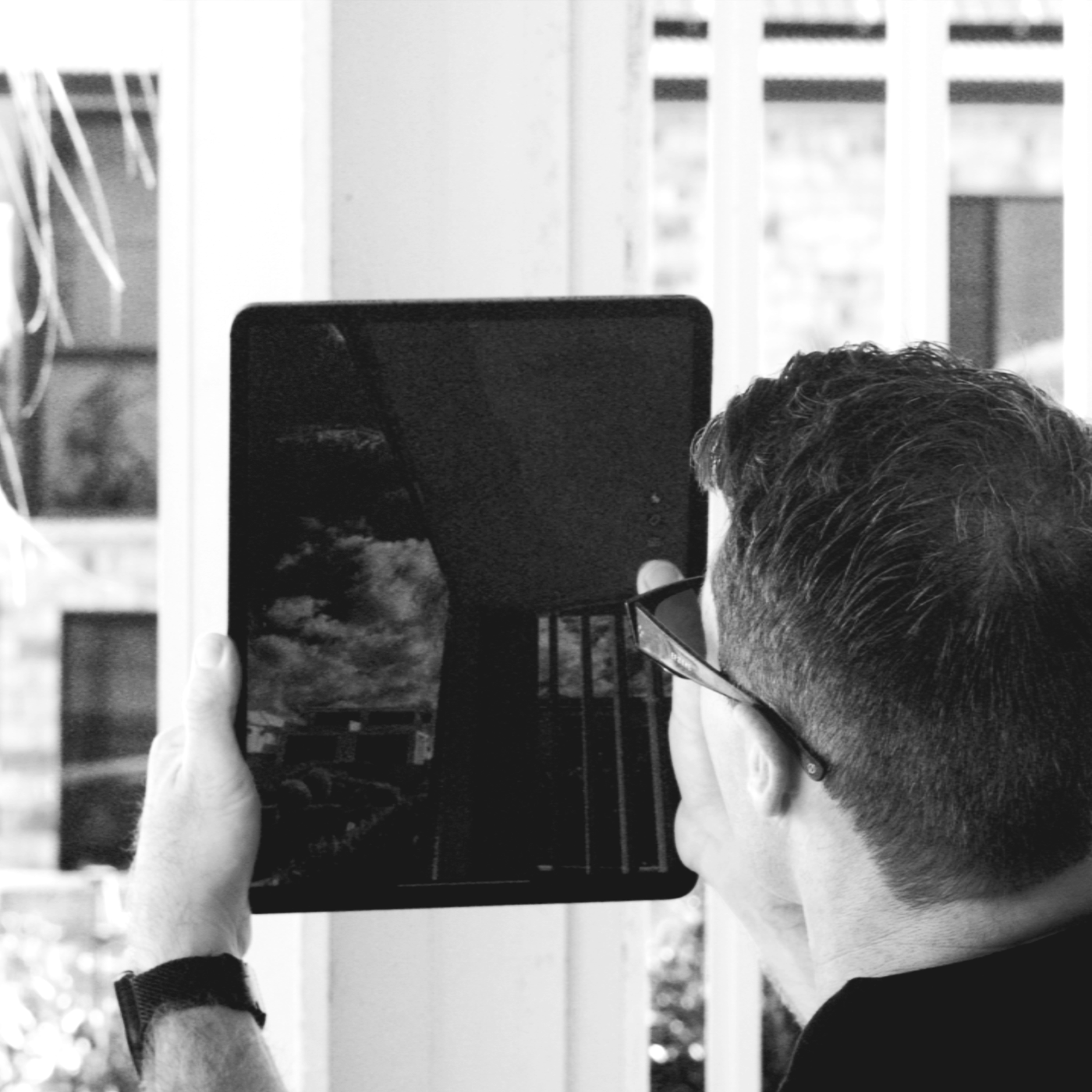 A person with glasses holds a tablet, capturing their reflection in the screen, which shows a cloudy sky. The person has short dark hair and is facing away from the camera.